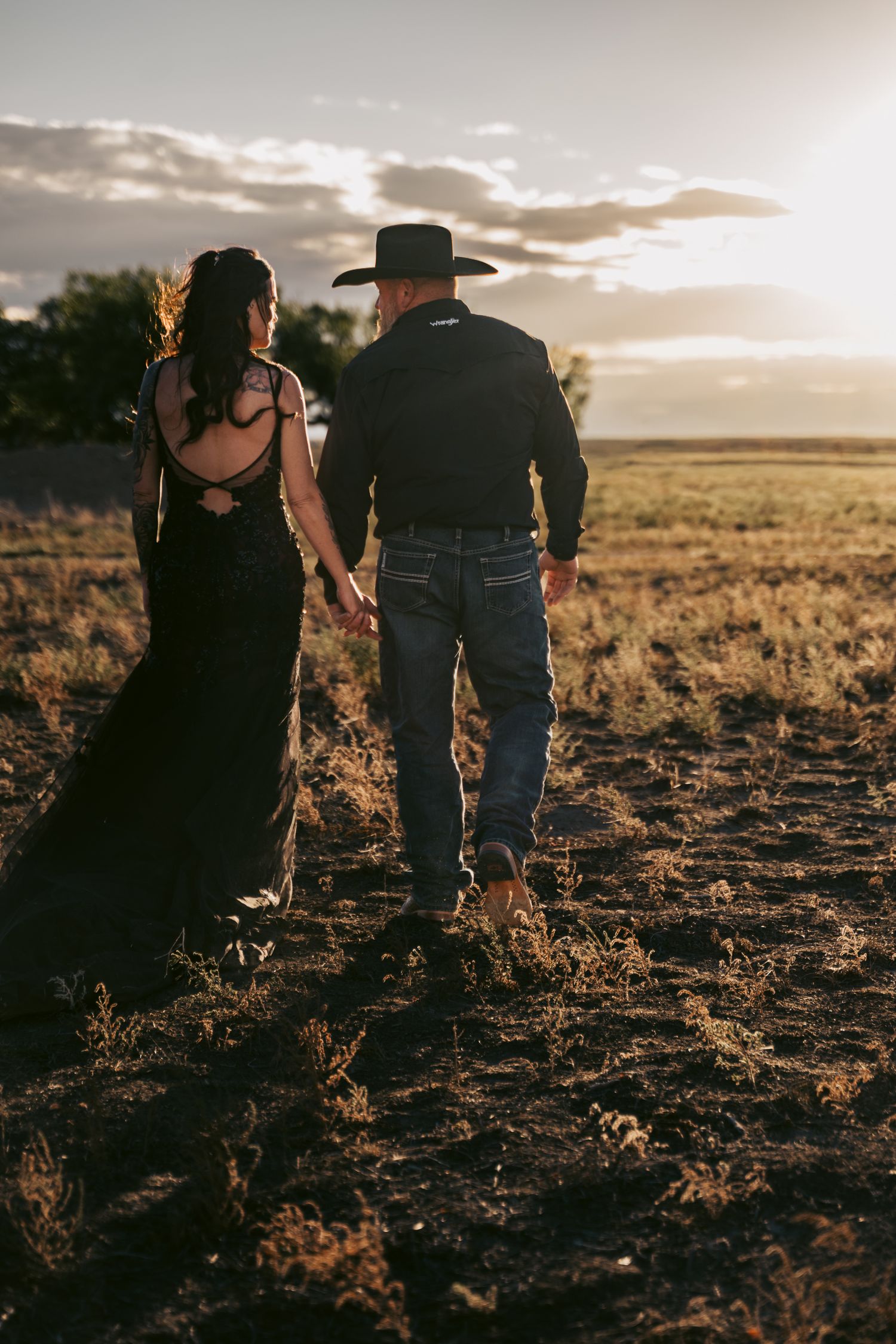 Couple in black western attire holding hands walking through a field at golden sunset hour.