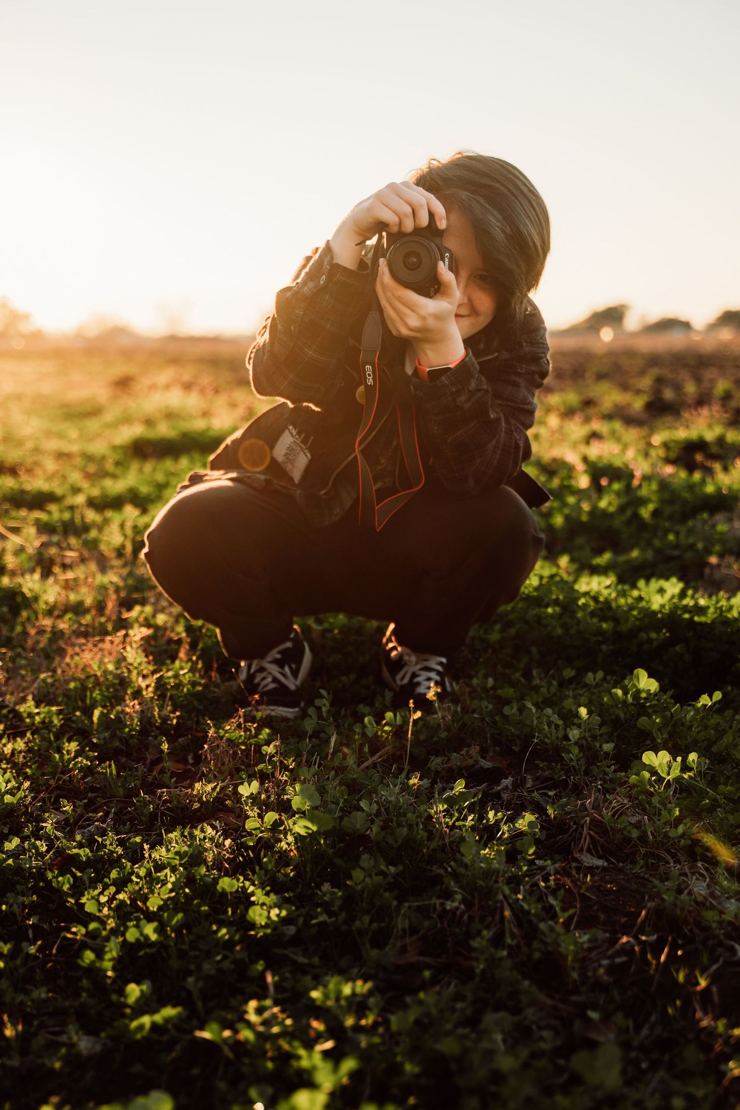 Photographer crouches in a field at golden hour taking photos among green foliage with sunlight streaming from behind.