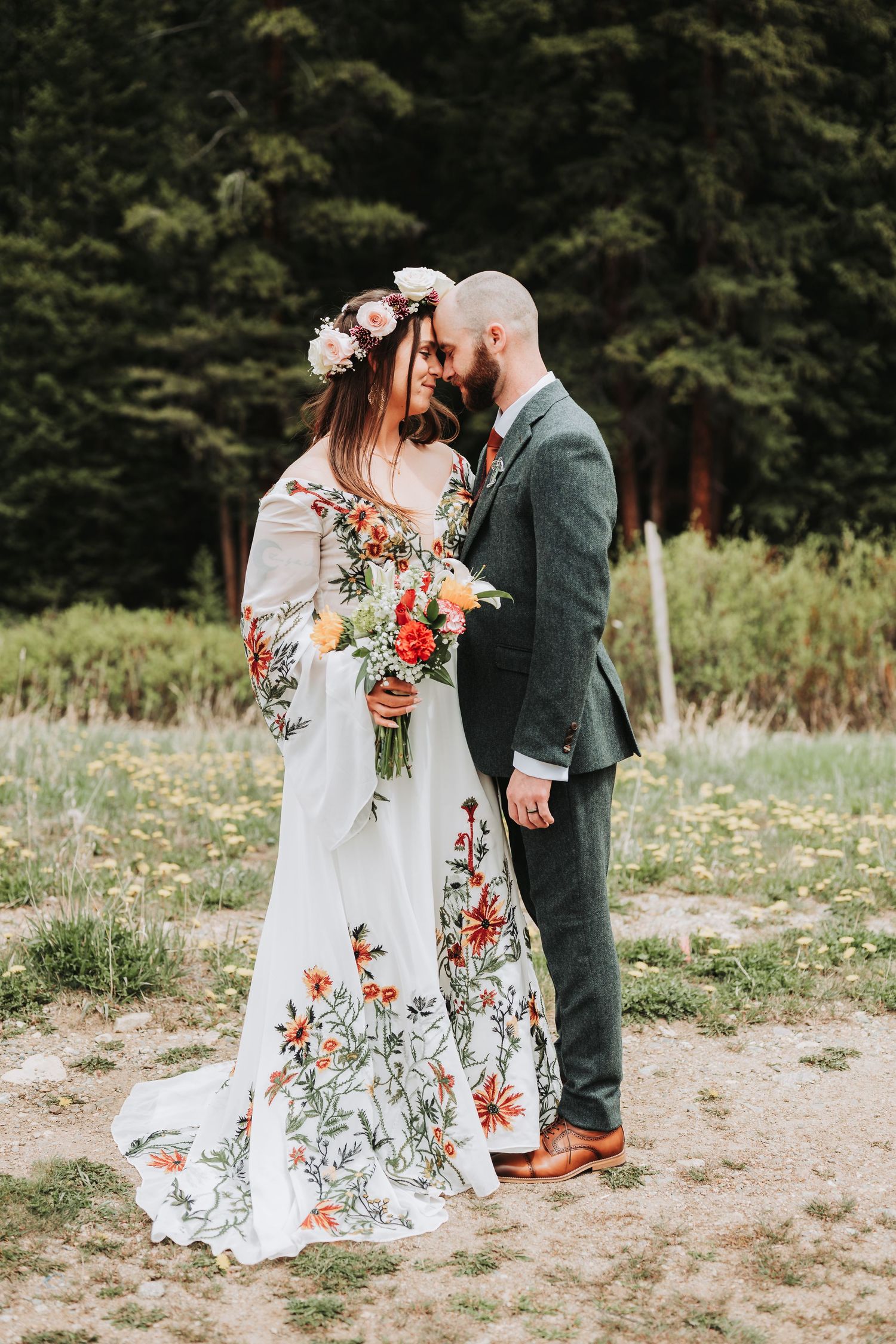 Bride in floral gown with flower crown and groom in gray suit share romantic moment outdoors in forest meadow.