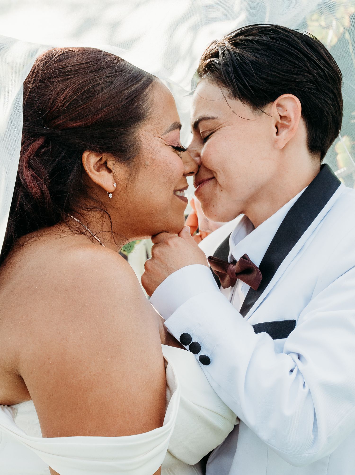 Two brides touch noses and smile under a sheer veil, one in a white strapless gown, the other in a white tuxedo jacket and burgundy bow tie.