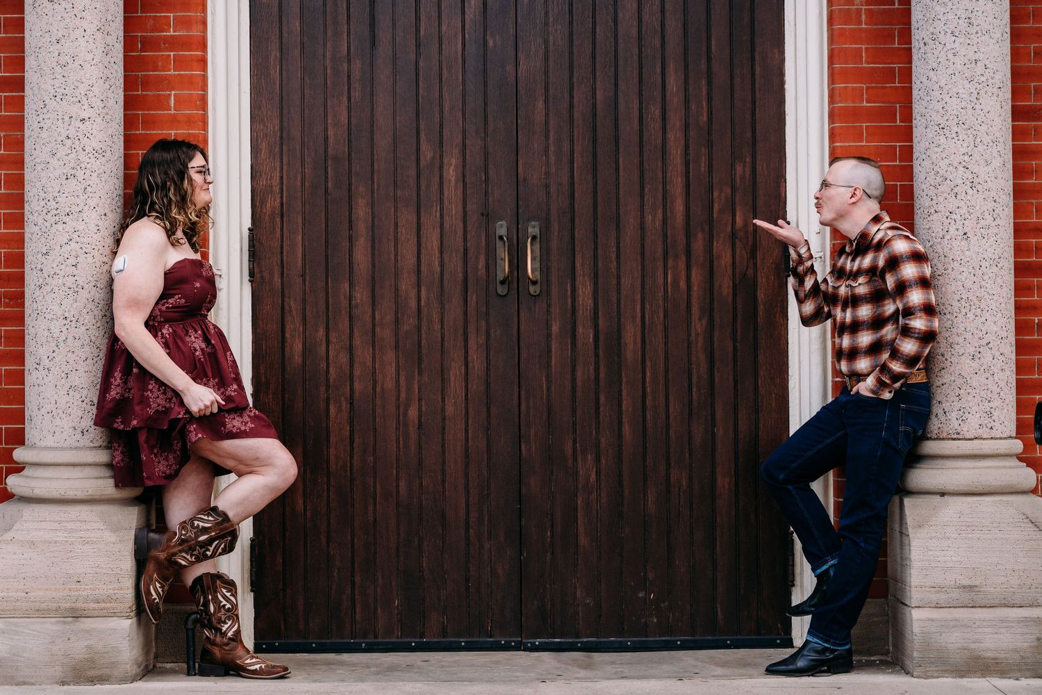 Couple posing playfully apart in front of a wooden door on brick building steps during an engagement photo session.