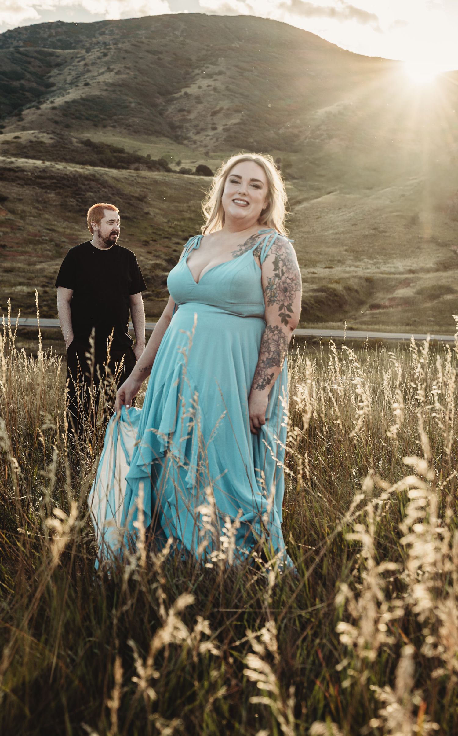 A couple poses in a grassy field at sunset with mountains in the background and sunlight creating a dreamy atmosphere.