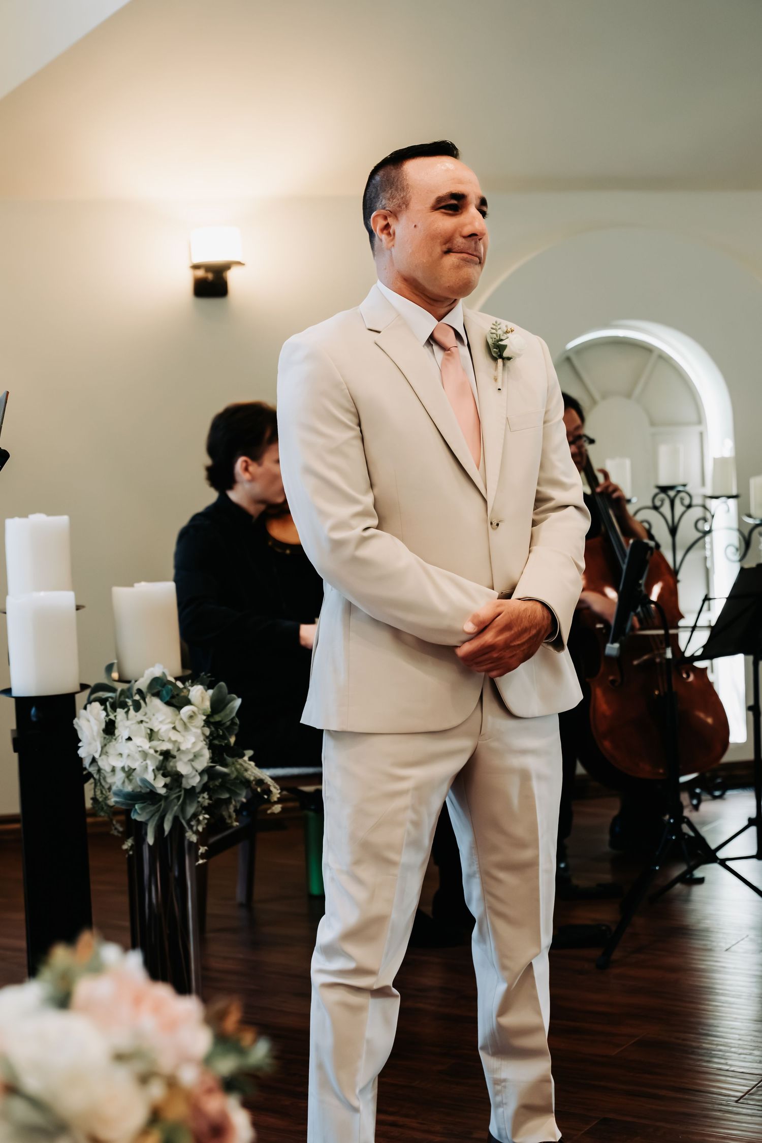 Groom in beige suit with pink tie smiles at altar during wedding ceremony with string musicians in background.