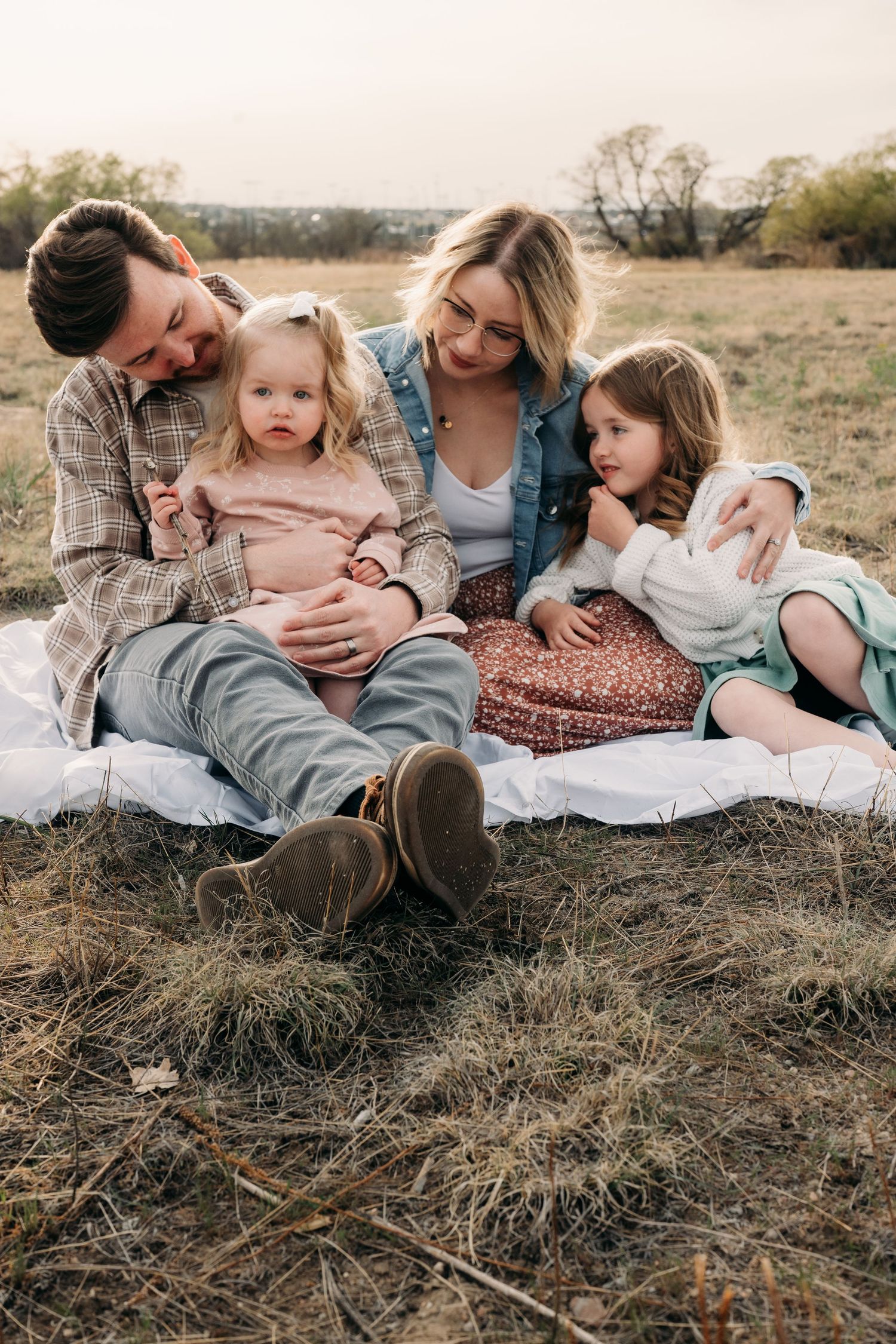 A family of four sits on a blanket in a field, with parents embracing two young daughters outdoors.