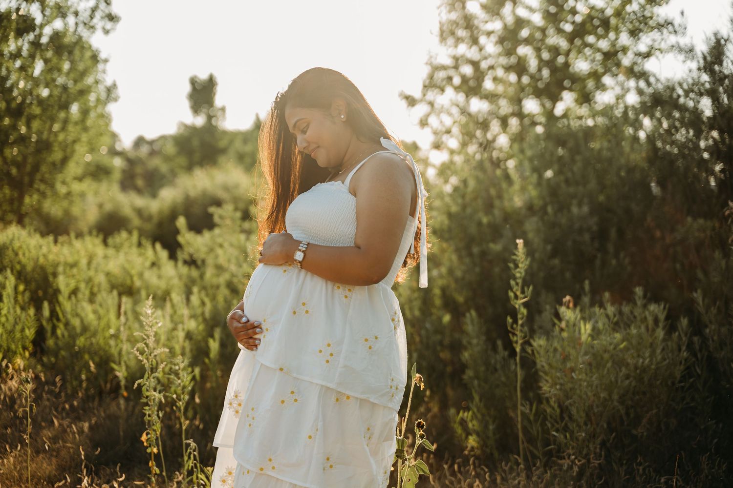 A couple in white outfits embraces during a golden hour photoshoot in a lush outdoor setting.