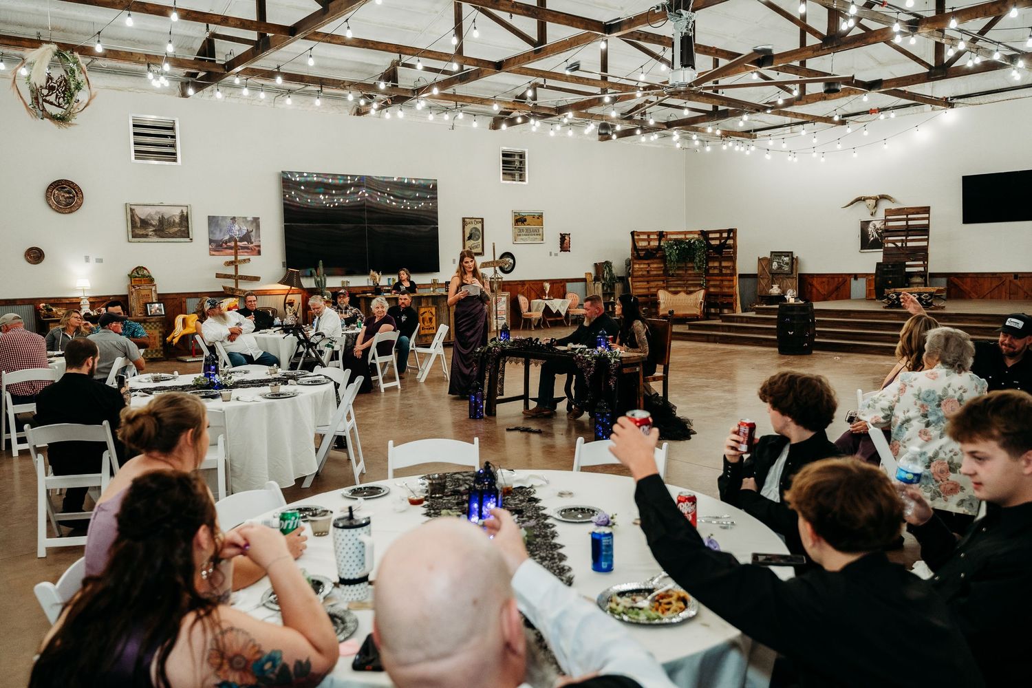 Guests seated at round tables during an indoor wedding reception in a rustic venue with string lights.