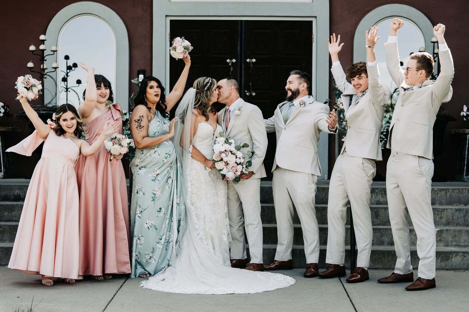Wedding party celebrates outdoors, bride and groom kiss while bridesmaids in pink and groomsmen in tan suits cheer.