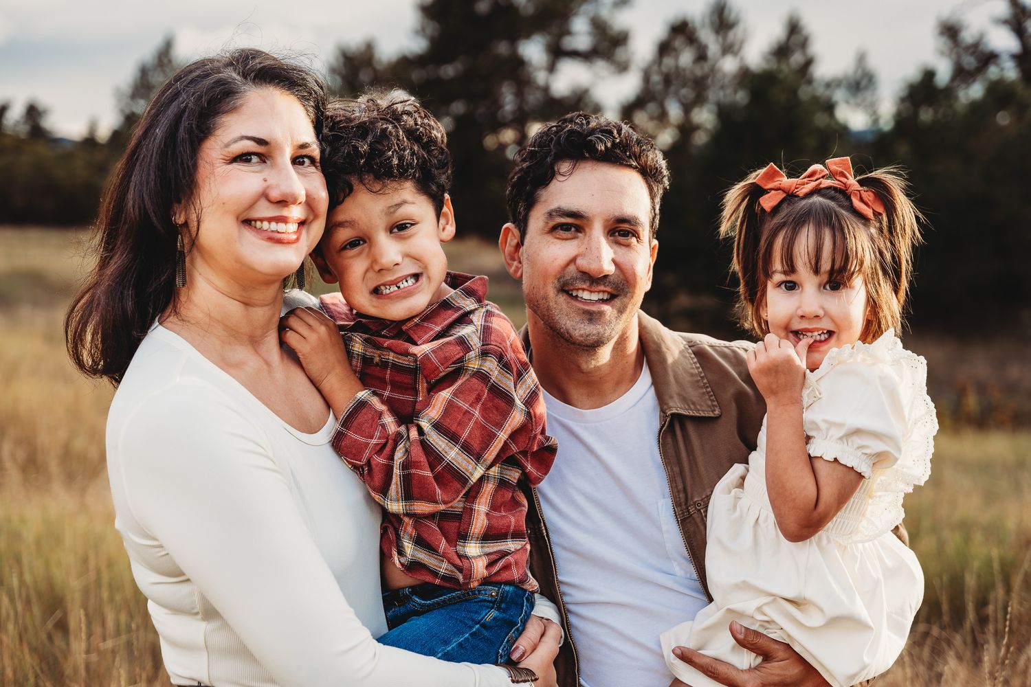 A family wearing coordinated casual outfits embraces and smiles together during an outdoor photoshoot at sunset.