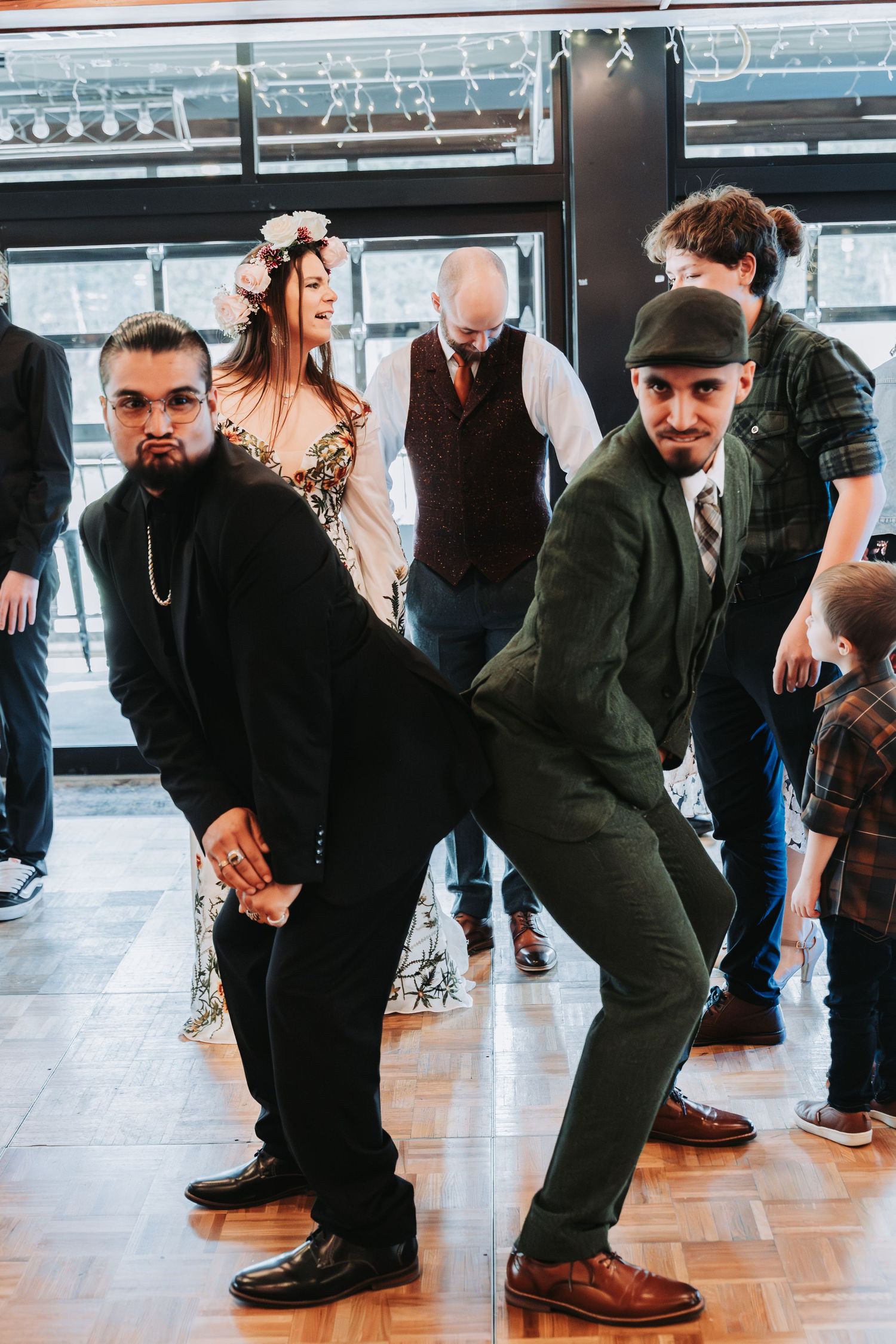 People dancing at a party, wearing stylish outfits including a floral headpiece and green suit, on a wooden dance floor.