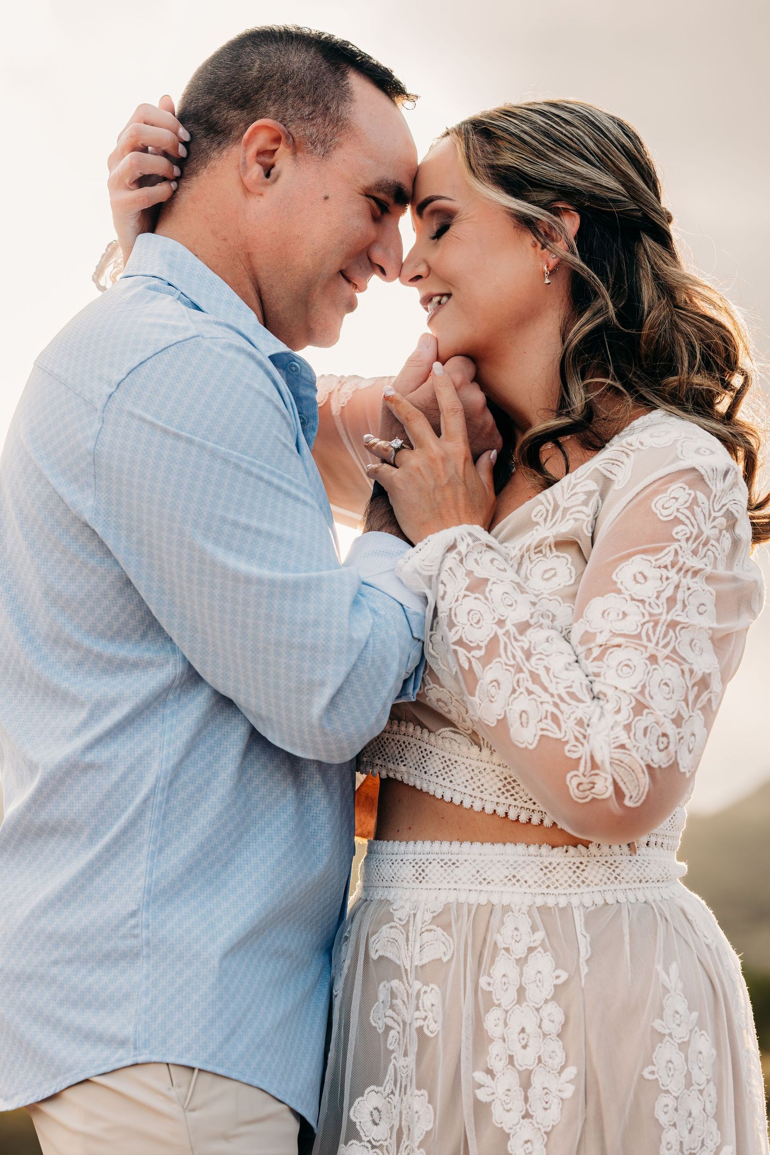 A couple in wedding attire smiles and touches foreheads tenderly, the bride wearing a lace dress and groom in a blue shirt.