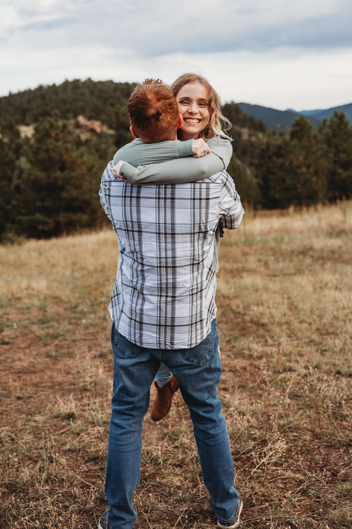 A couple embraces in a field with mountains and dry grass in the background.