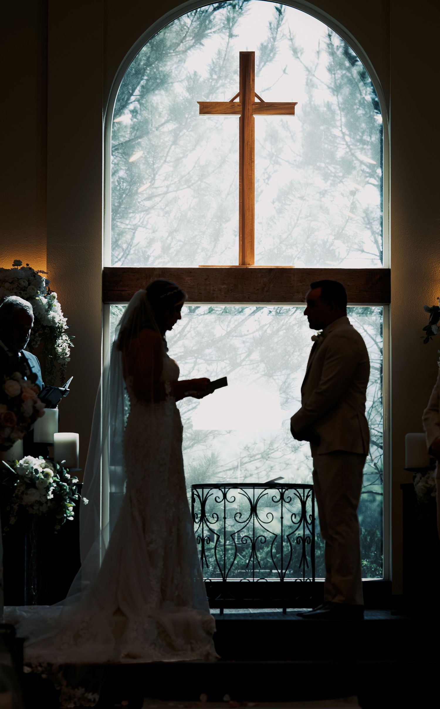 Silhouette of bride and groom exchanging vows before a wooden cross in a chapel window during wedding ceremony.