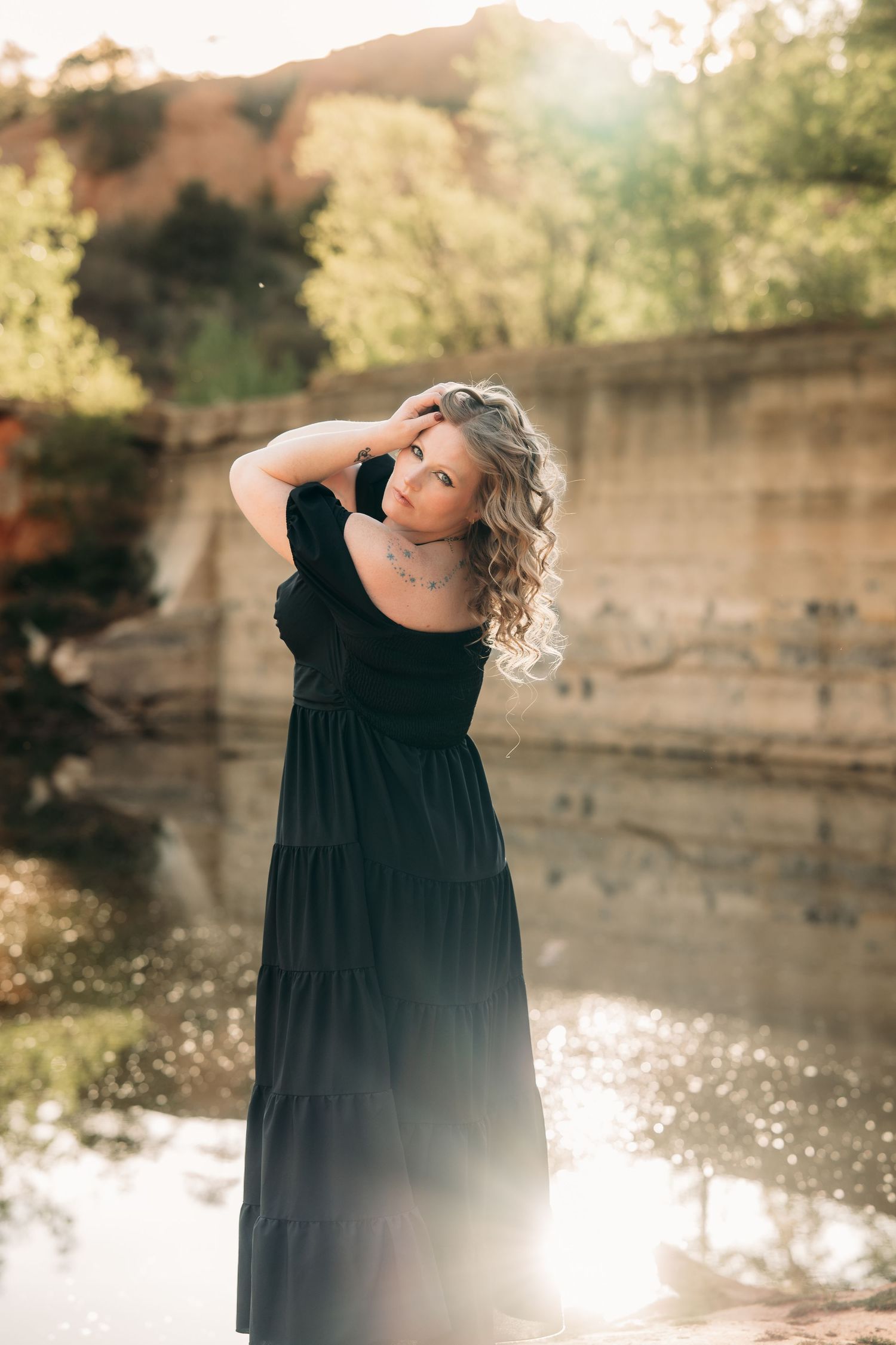 Woman in black maxi dress poses near sunlit rocky pond surrounded by lush green trees in golden hour light.