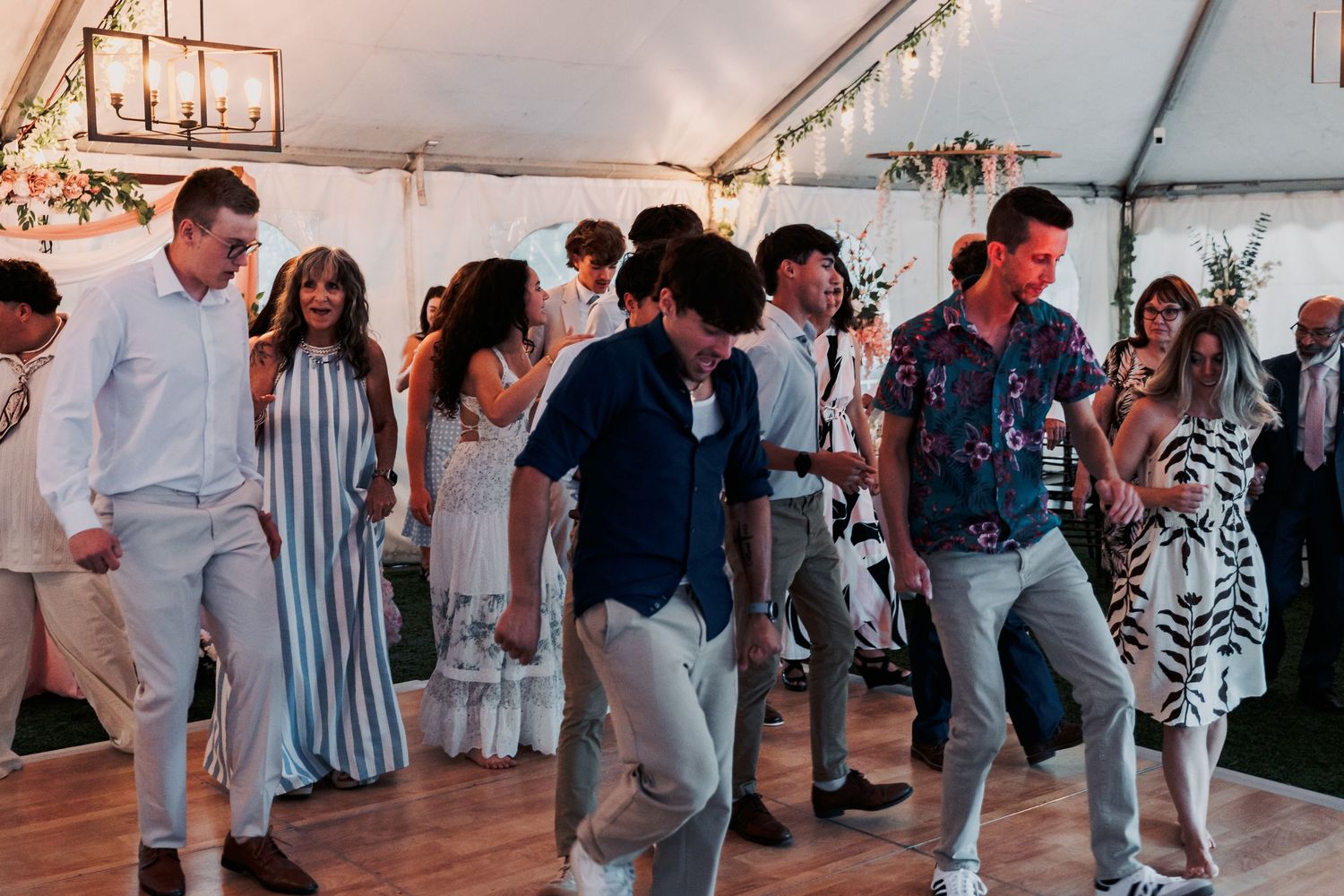 Guests dancing energetically at an outdoor wedding reception under a white tent with string lights.