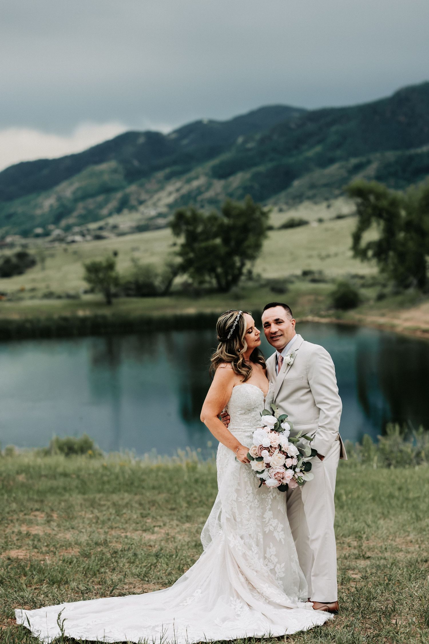 Bride and groom pose by a mountain lake, she in lace gown with bouquet, he in tan suit, green hills behind.