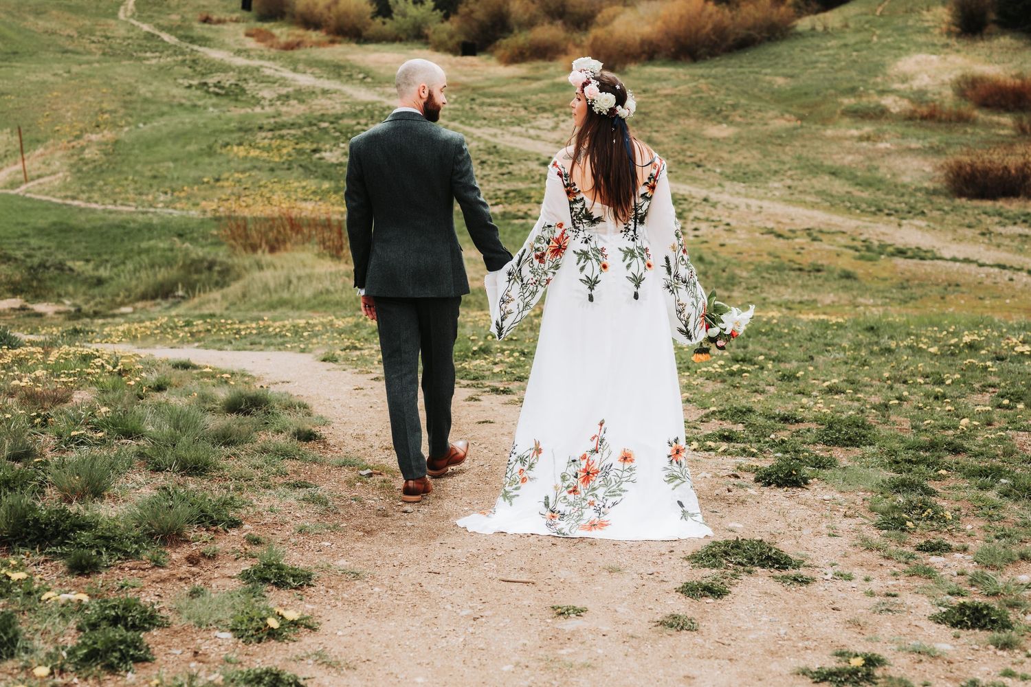 A bride in a floral embroidered white gown and groom in dark suit walk hand in hand along a rustic dirt path.