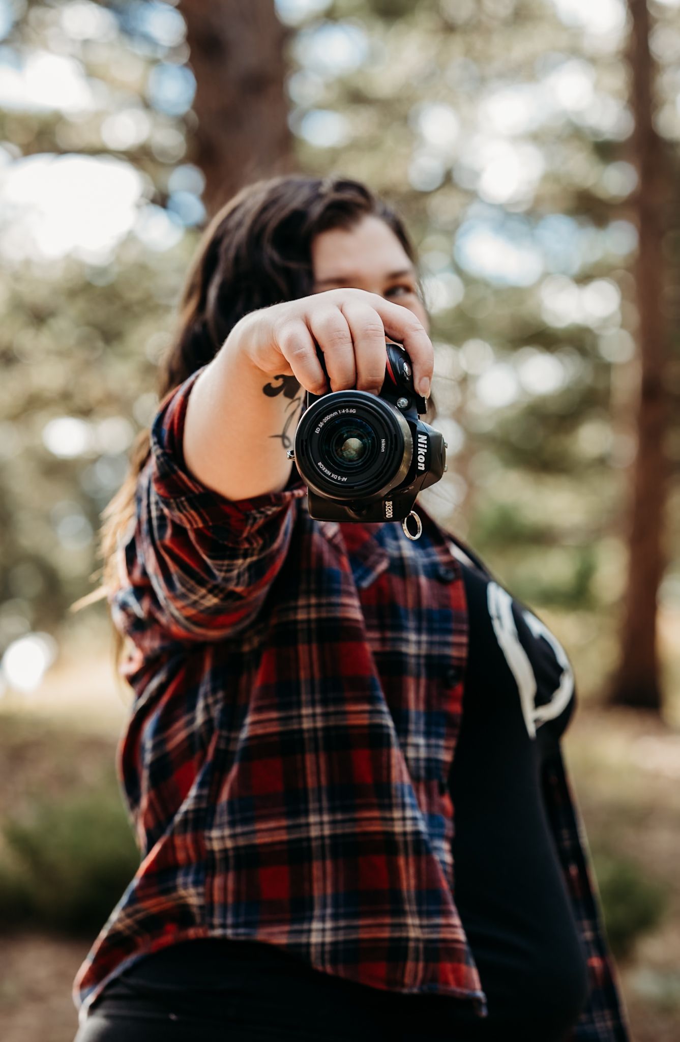 A young woman in a plaid shirt holds a Nikon camera up to photograph outdoors in a forest setting.