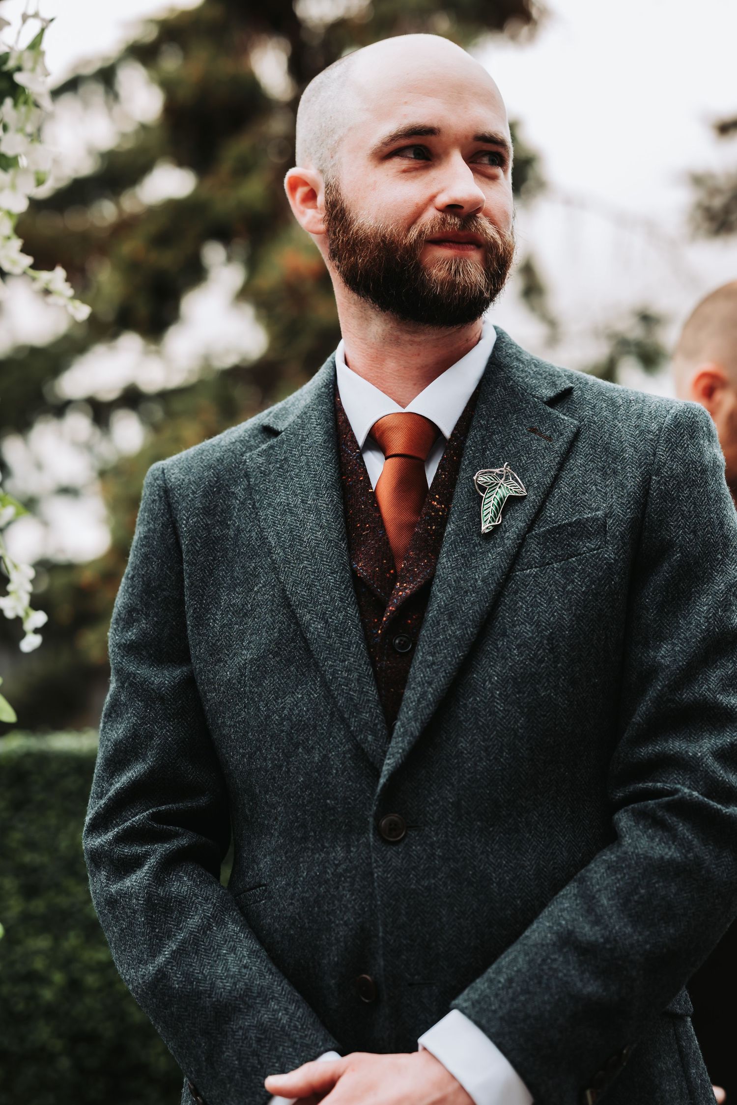 Bald groom with beard wearing gray tweed suit, rust tie, and brooch boutonniere at outdoor wedding ceremony.