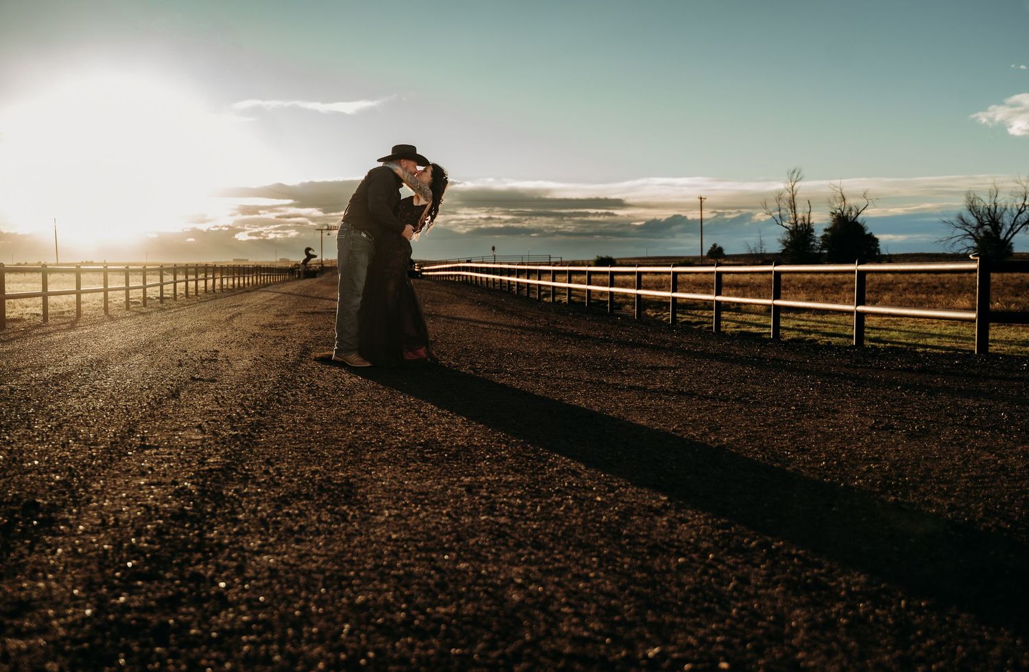 Couple sharing a romantic kiss on a rural road at sunset, with wooden fences and golden light in background.
