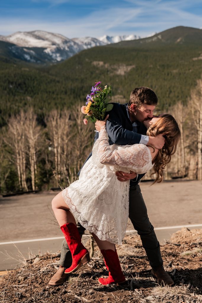 A couple in wedding attire share a romantic dip pose against a backdrop of snow-capped mountains.