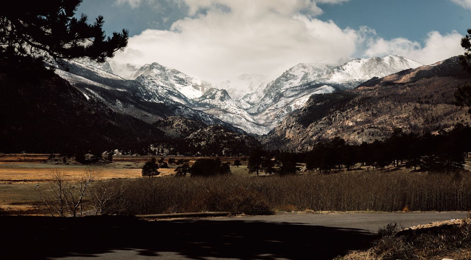 Snow-capped mountain range towers over a scenic valley with bare trees and open fields under a cloudy sky.