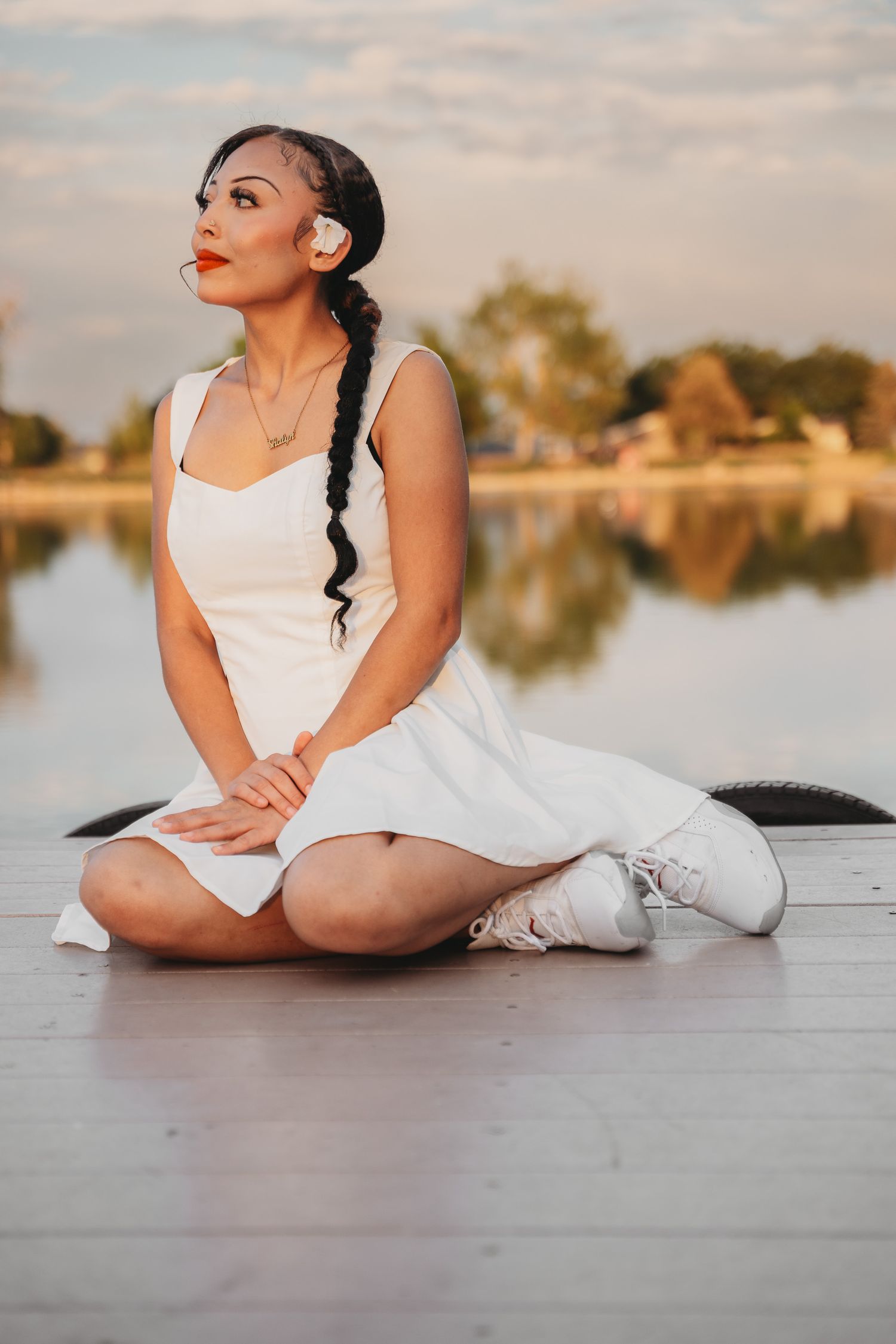 Woman in white dress and sneakers sitting on a dock by a lake at sunset with a braided hairstyle.
