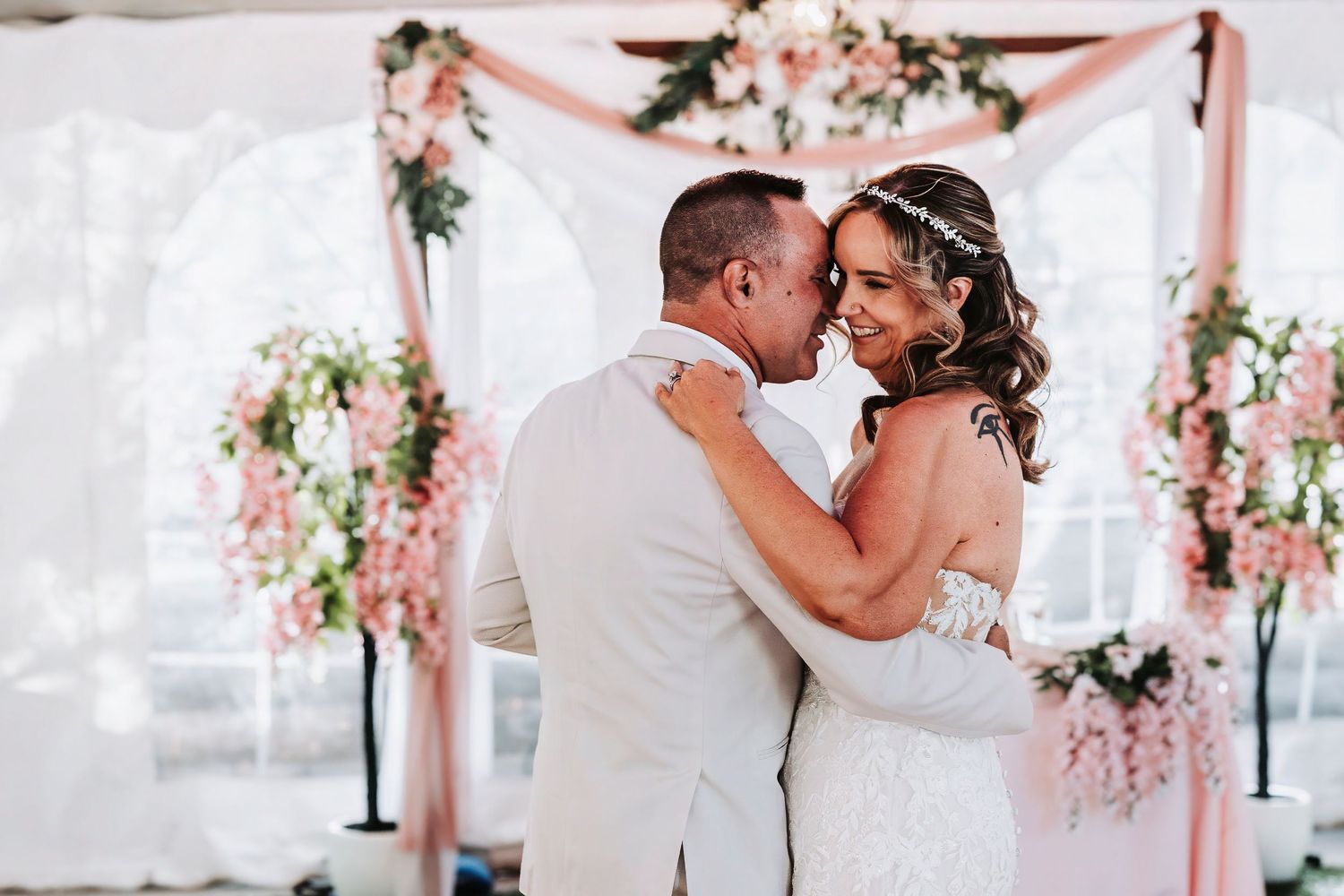 Bride and groom share a romantic moment at floral wedding altar with pink blooms and white draping.