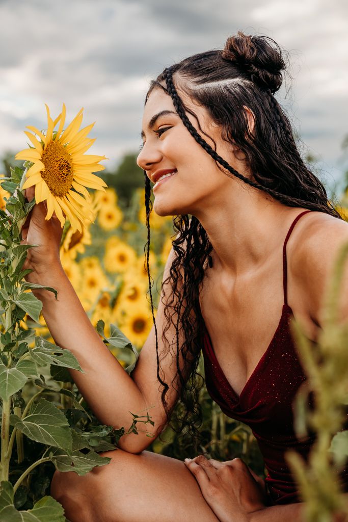 Dark-haired figure poses joyfully in a vibrant sunflower field on a cloudy summer day.