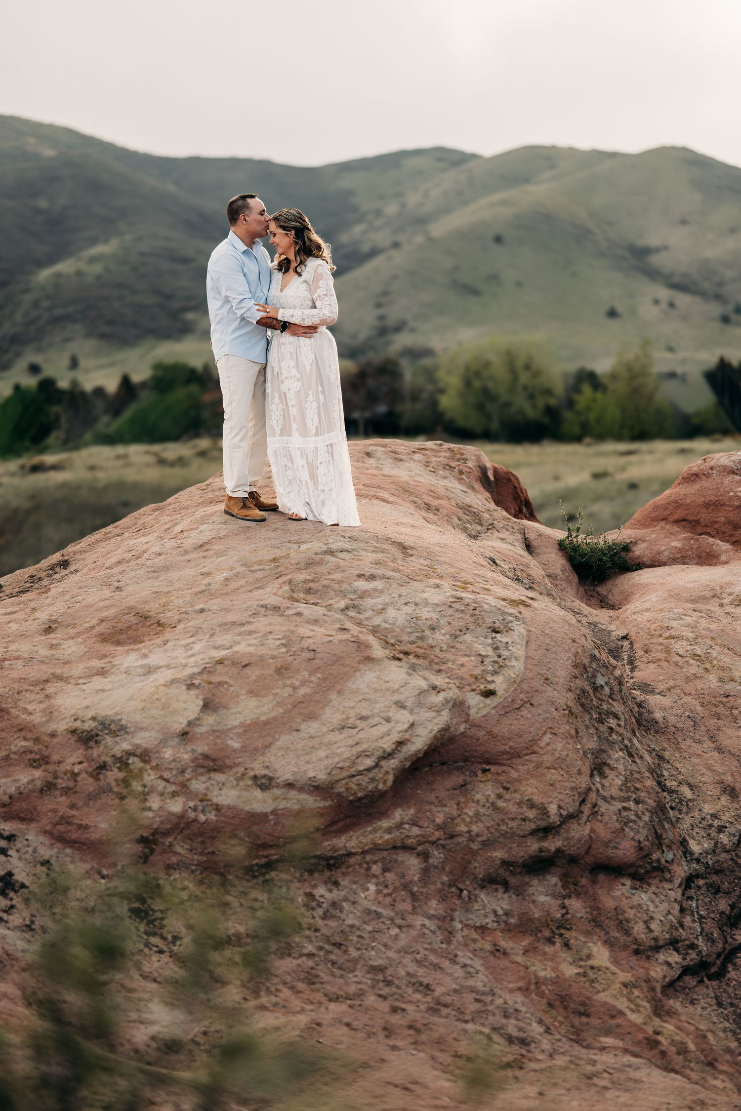 Couple embracing on red rock formation with green mountain backdrop during outdoor engagement photo session.