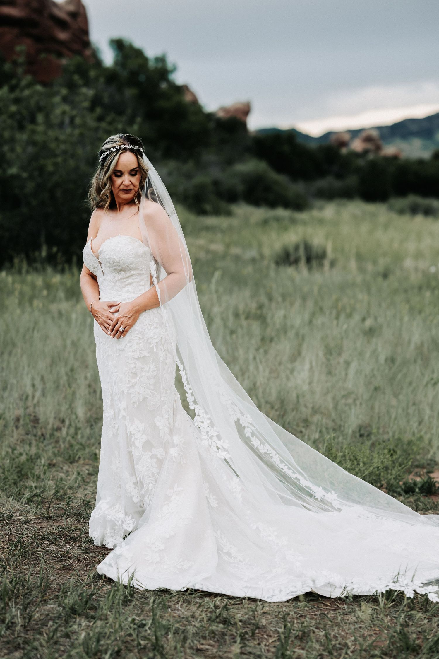Bride in lace mermaid gown with cathedral veil stands in green meadow with red rock formations backdrop.
