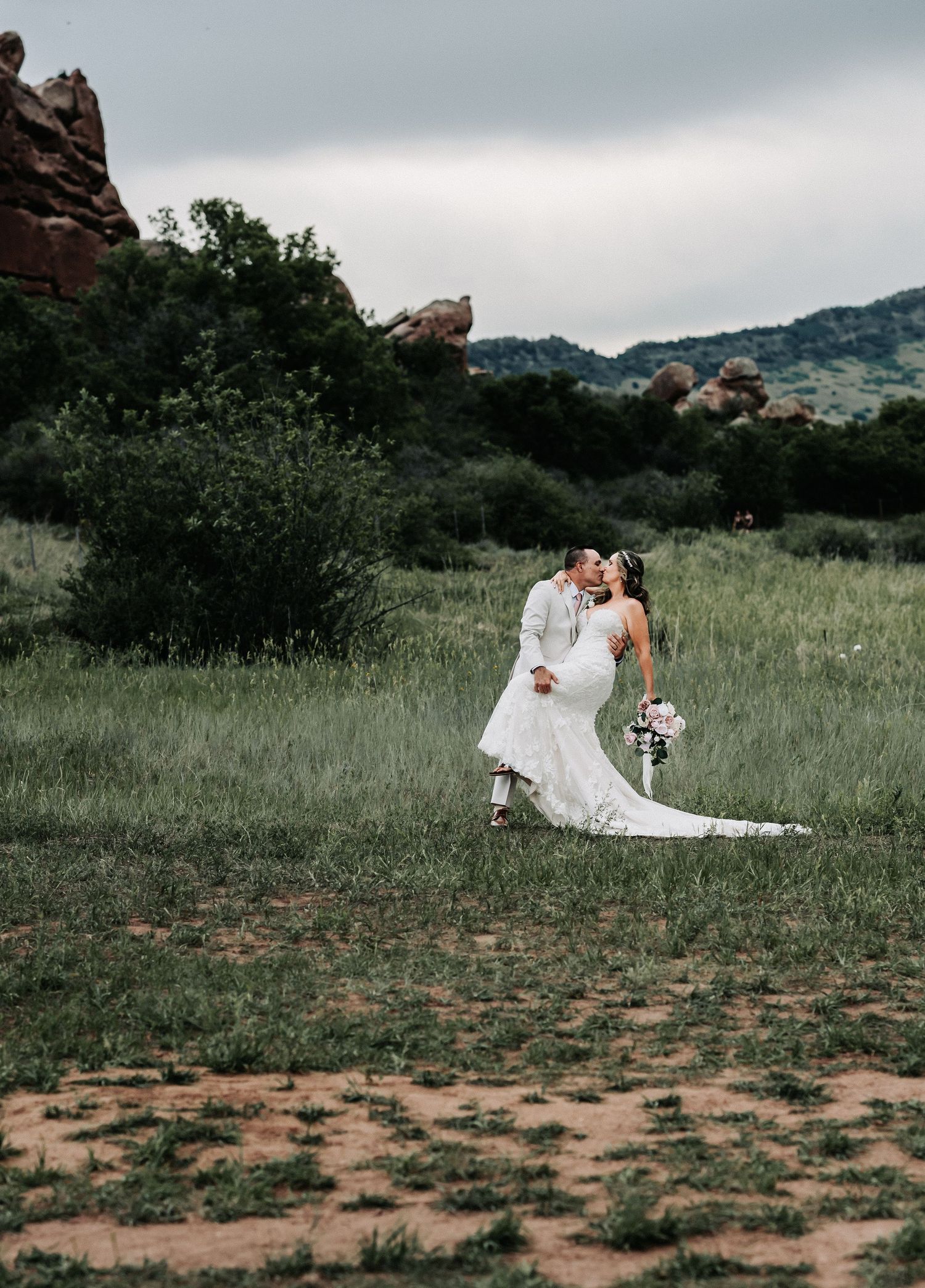 A bride and groom share a romantic dip kiss in a lush green meadow with red rock formations under a cloudy sky.