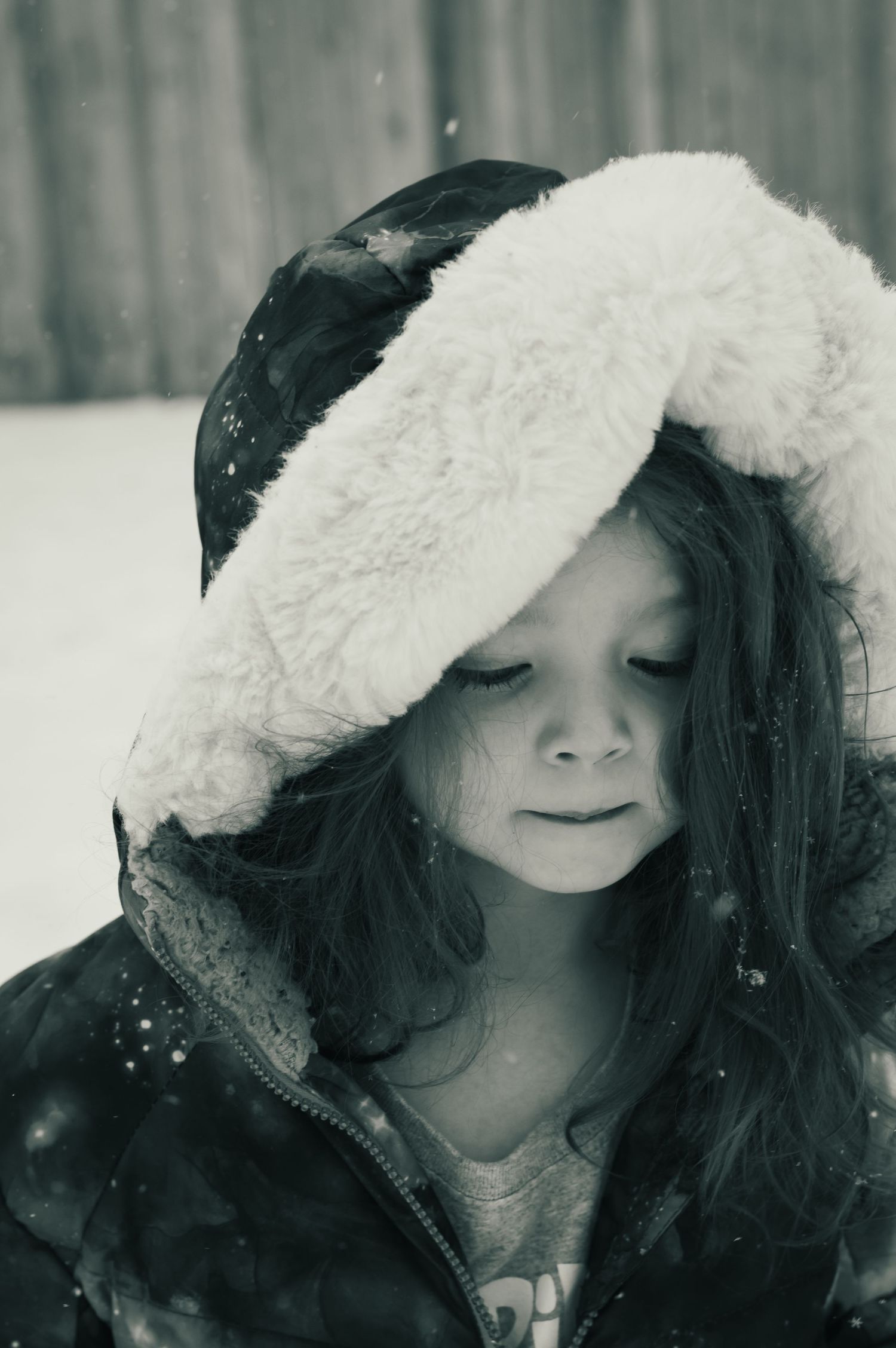 A winter portrait in black and white showing someone wearing a fluffy white hooded coat while snow falls around them.