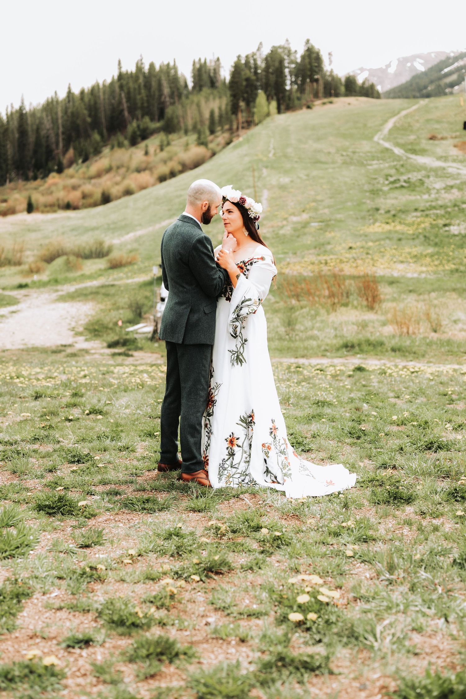 Bride in floral gown and groom in dark suit share romantic moment on green hillside with pine trees backdrop.