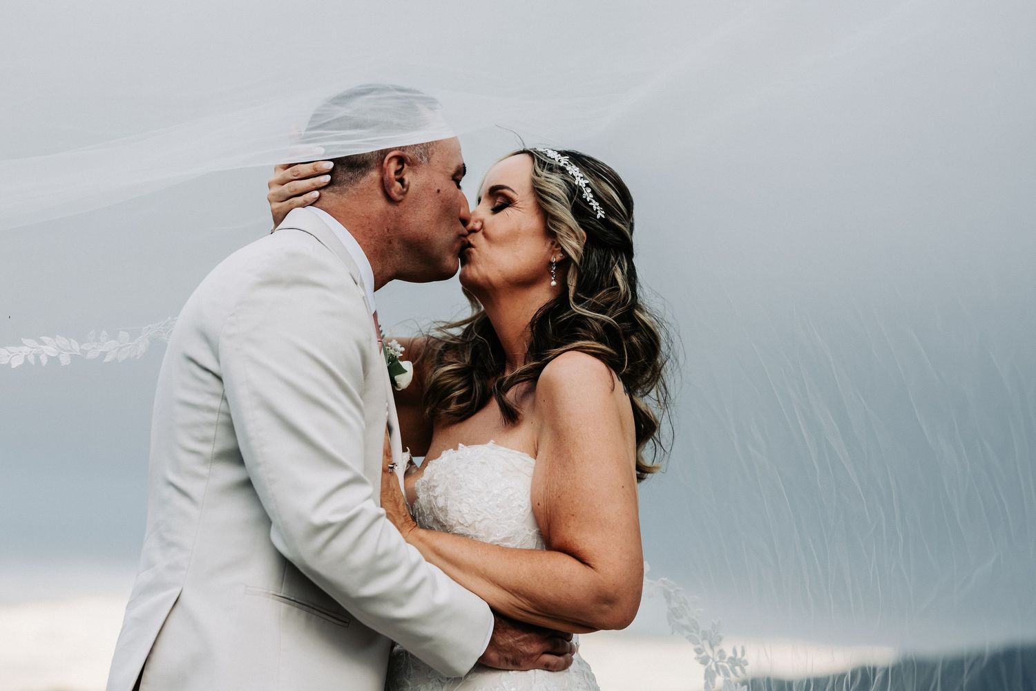 A couple shares a romantic kiss in the rain on their wedding day, with the groom in a white suit and bride in a strapless gown.