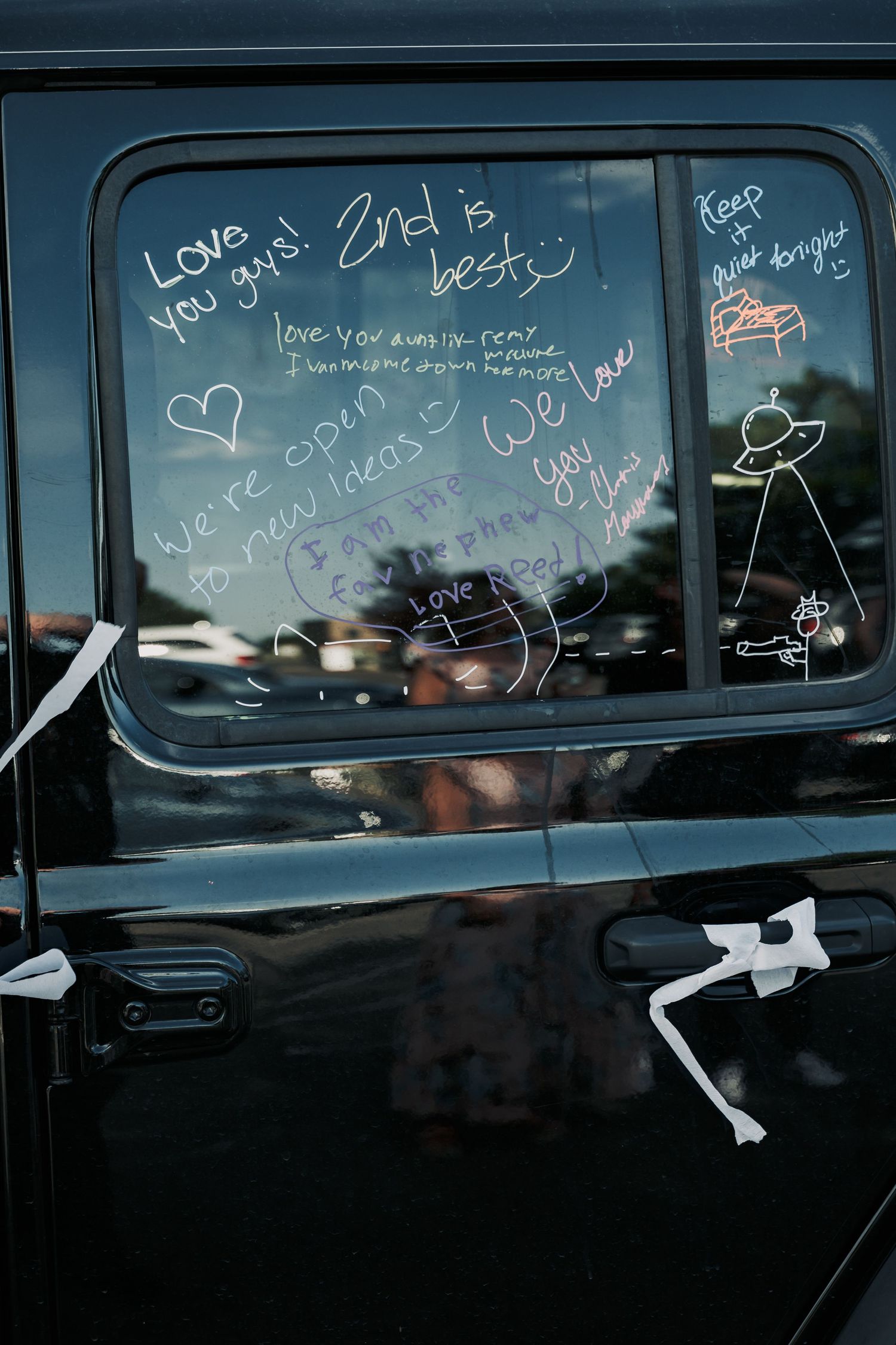 Black vehicle window decorated with white marker wedding congratulations messages and stick figure drawings.
