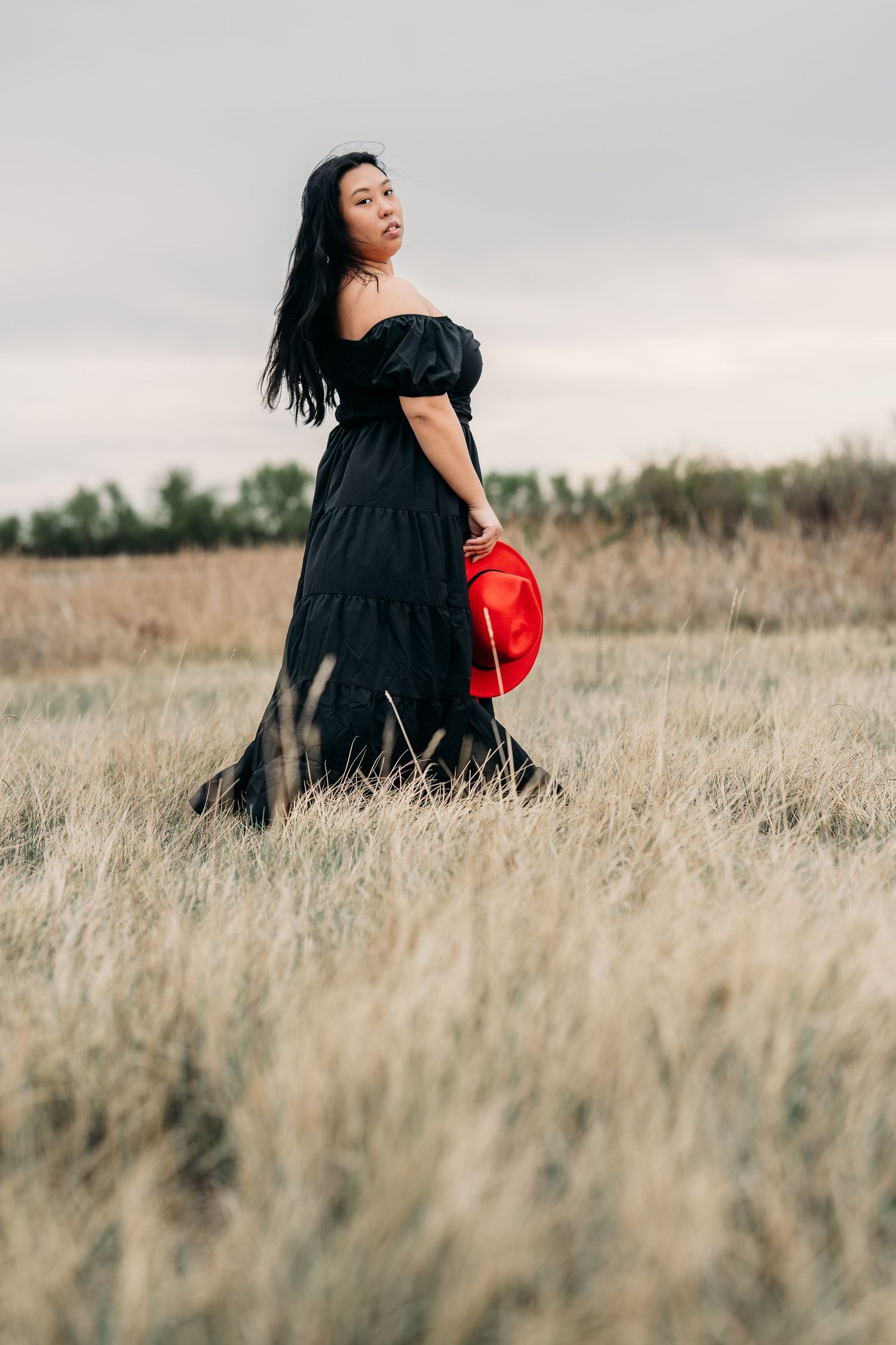 Woman in black off-shoulder dress holding red hat walks through dry grass field under overcast sky.