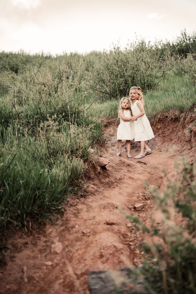 Two children in white dresses walk along a rustic dirt path surrounded by tall green grass and vegetation.