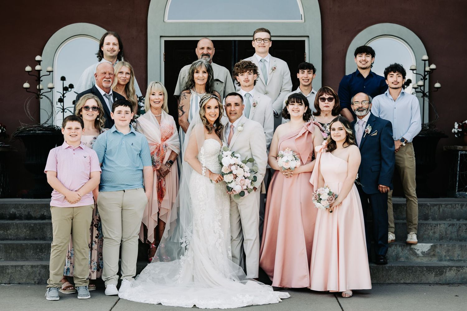 Wedding party poses on church steps, bride in white gown with groom and family in formal attire.