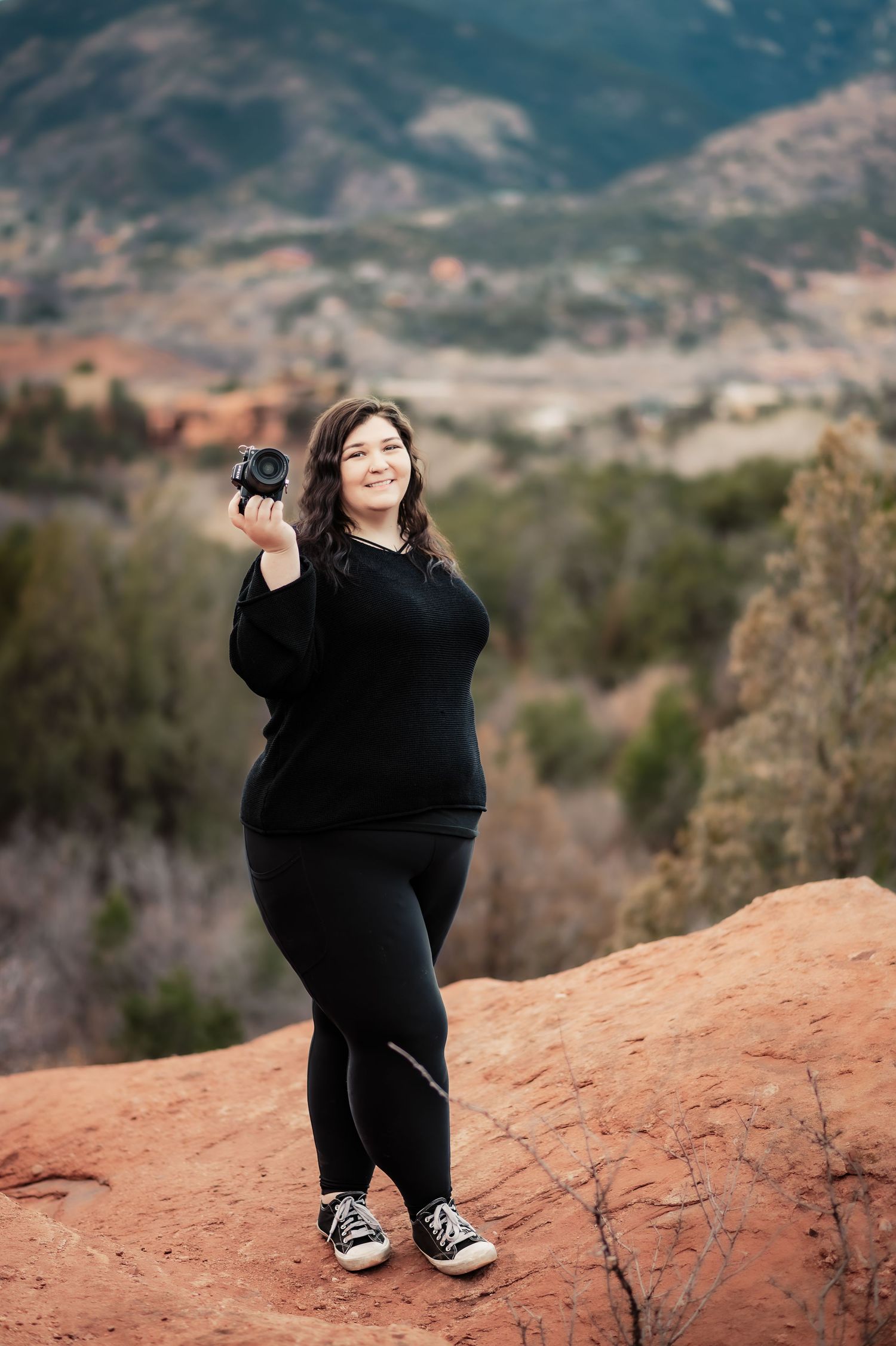 Woman in black outfit holding camera on red rocks with mountain landscape backdrop.