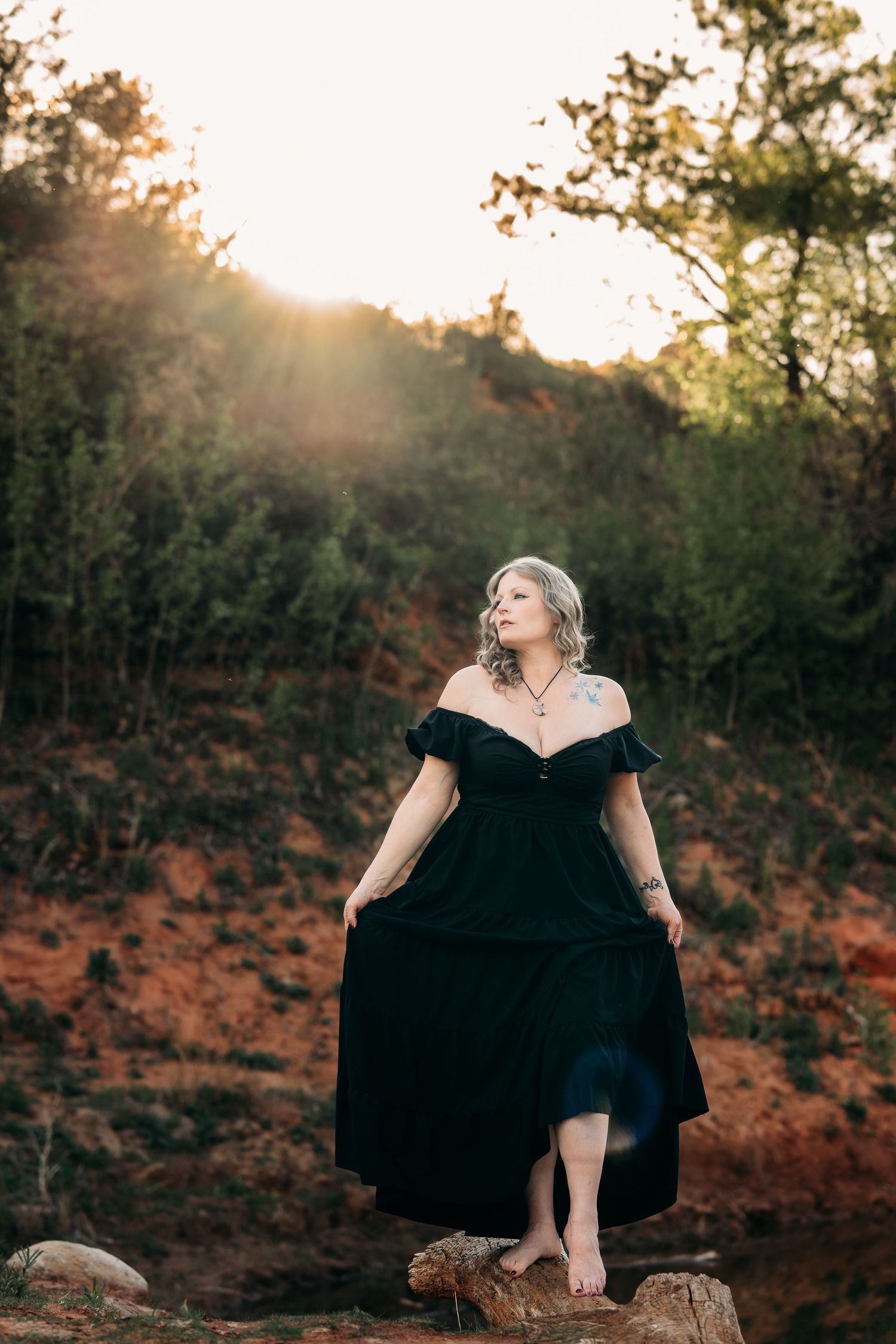 Woman in black off-shoulder gown stands barefoot on rocks at golden hour in a wooded outdoor setting.