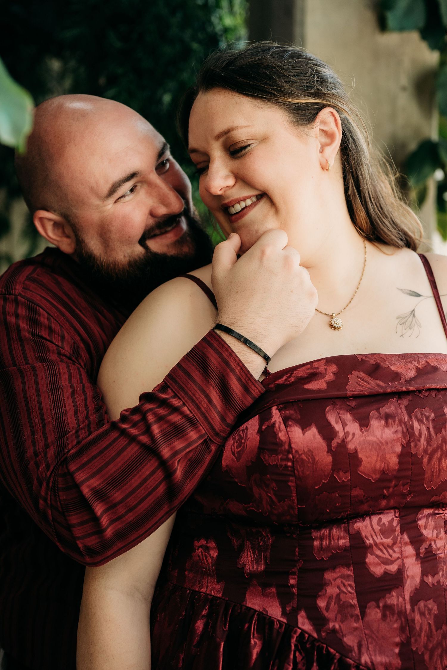 A bald man in red plaid embraces a smiling woman in a burgundy dress, sharing a tender romantic moment outdoors.
