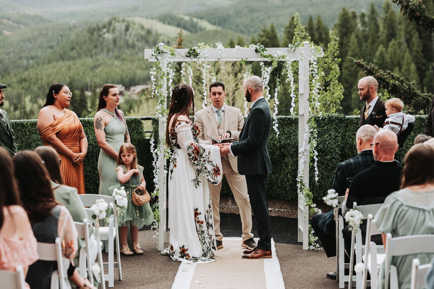 Outdoor wedding ceremony on a mountain terrace with floral arch, bride and groom exchanging vows before guests.