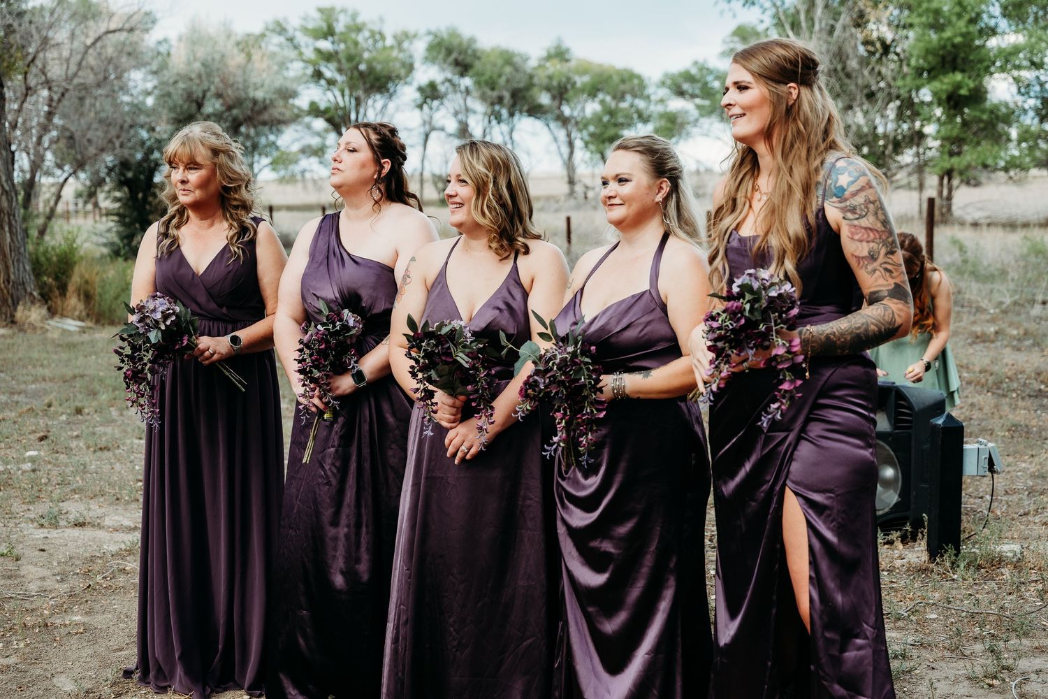 Four bridesmaids in purple satin dresses holding dark floral bouquets stand outdoors at a rustic wedding venue.