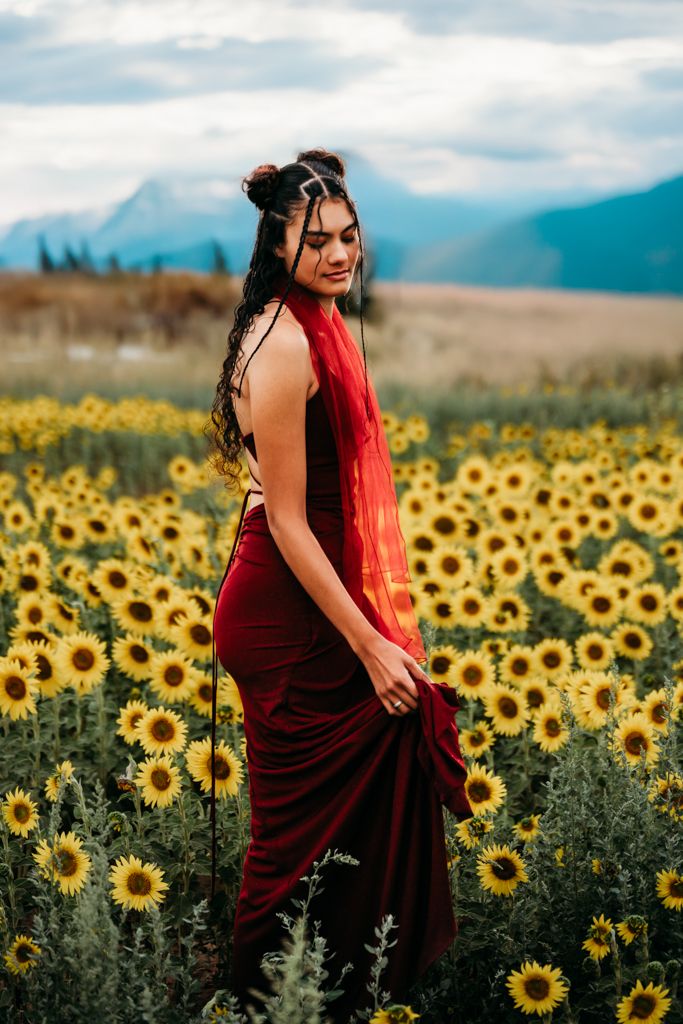 A figure in a burgundy dress twirls joyfully through a vibrant field of blooming sunflowers with mountains in the distance.