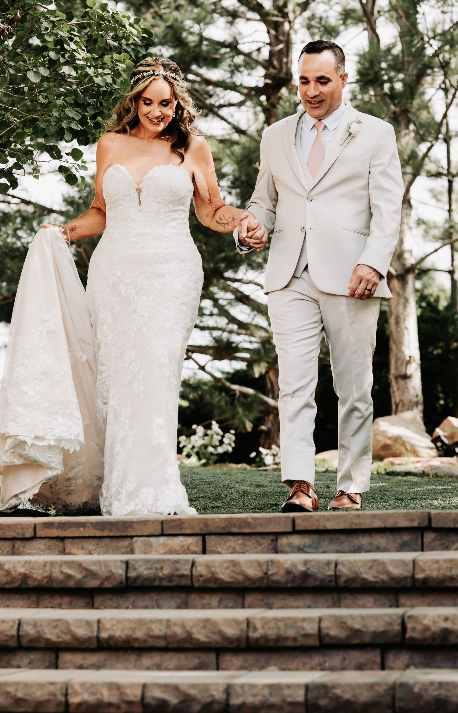 A bride in a white lace gown and groom in a tan suit hold hands while descending stone steps outdoors.