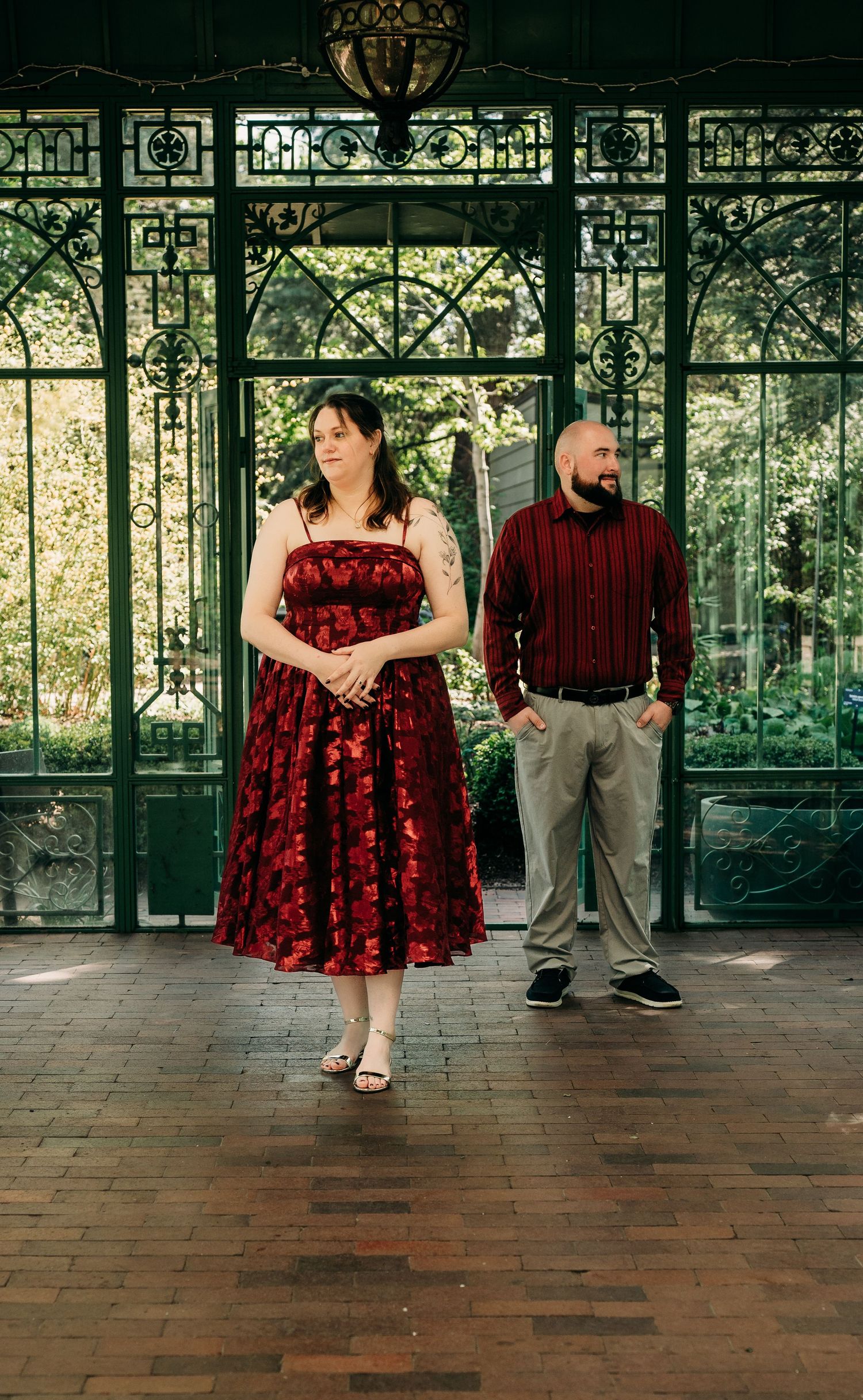 Couple in matching red and burgundy outfits posing before ornate green iron greenhouse doors on brick floor.