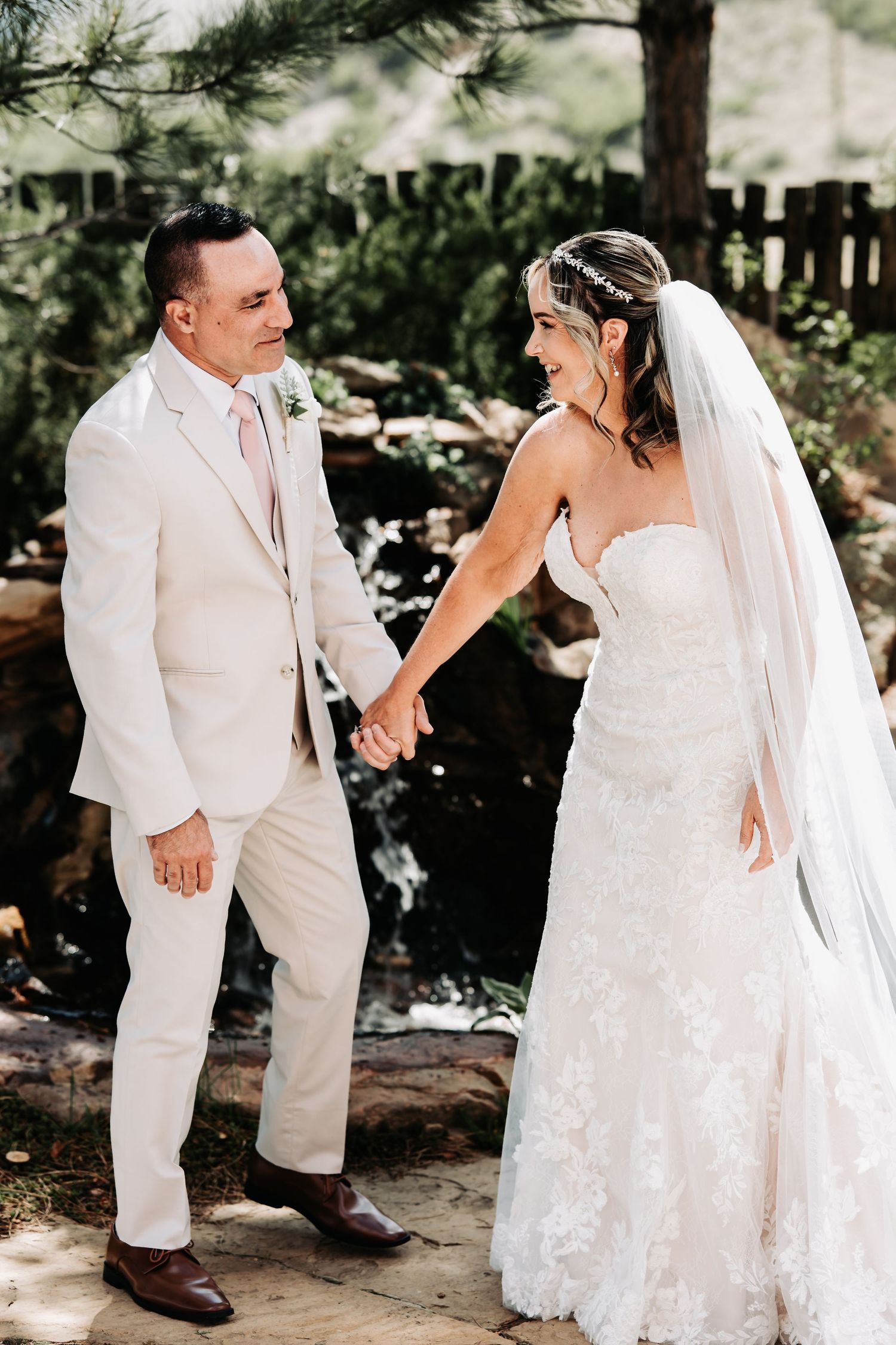 Bride in lace gown and veil holds hands with groom in cream suit, smiling outdoors near a garden waterfall.
