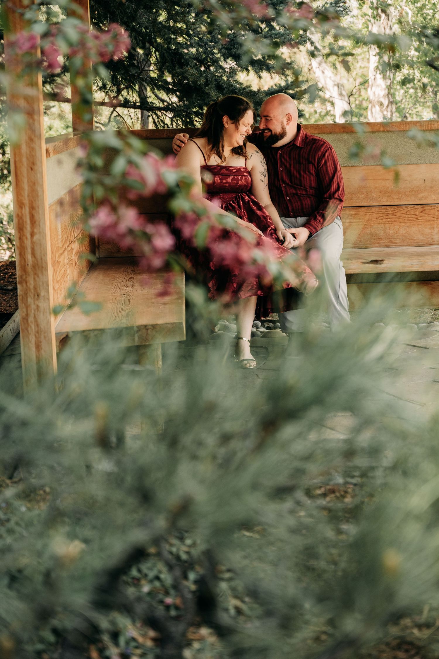 Couple in matching burgundy outfits sitting close together on a wooden bench surrounded by lush greenery and pink flowers.