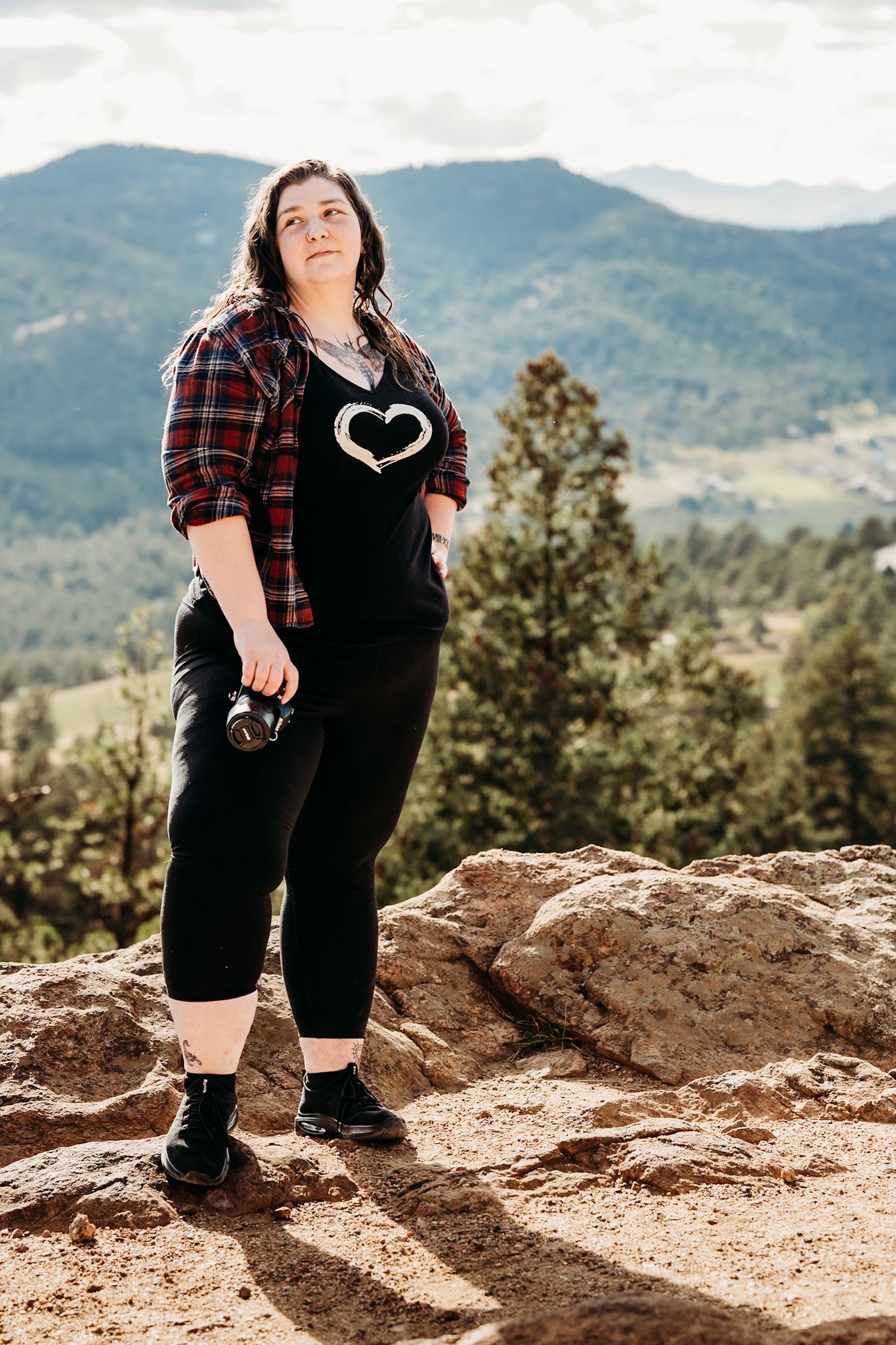 Woman in plaid shirt and black overalls stands on rocky overlook with mountain forest views behind her.