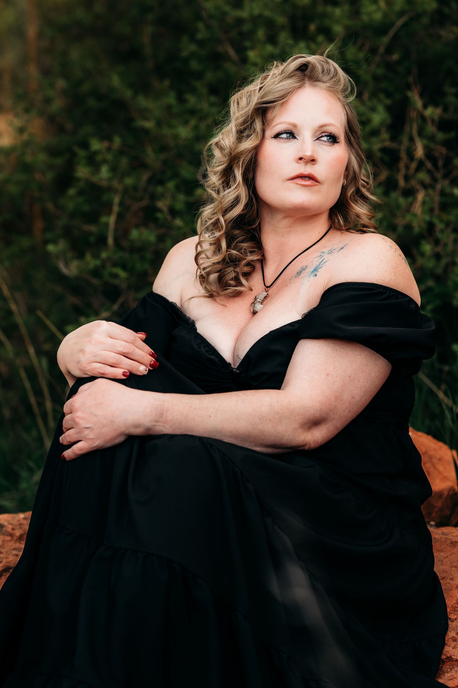 Woman in black off-shoulder gown sitting outdoors among green foliage, wearing necklace with tattoo visible.